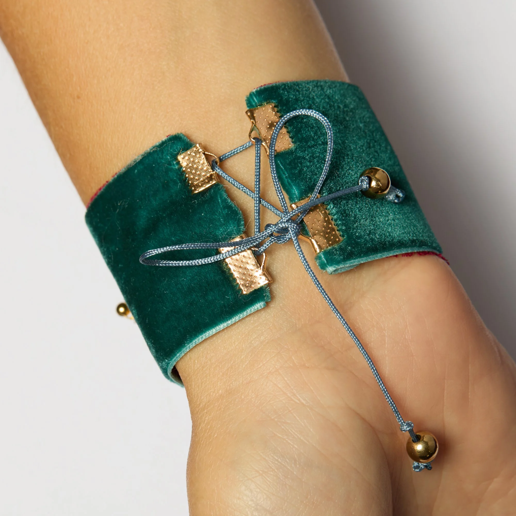Close-up of a person's wrist wearing a green velvet bracelet with gold and silver charms and a blue string bracelet with gold beads.