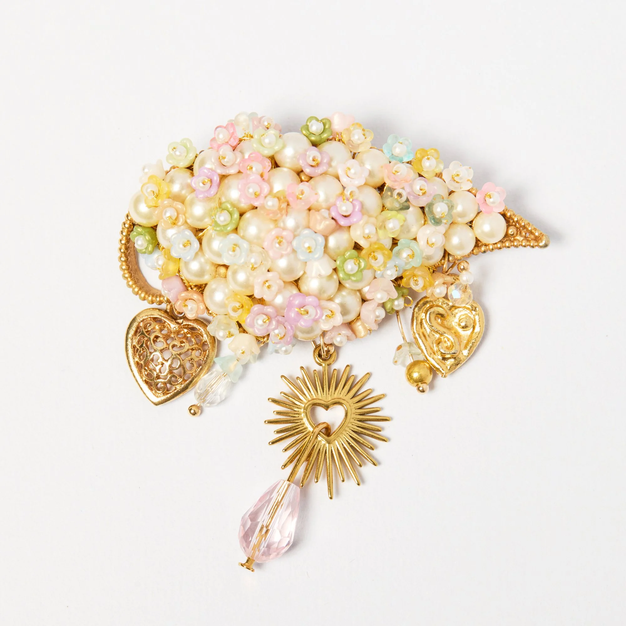 A decorative brooch with pearl and floral embellishments, gold accents, a pink crystal drop, and a sun-shaped charm, against a white background.