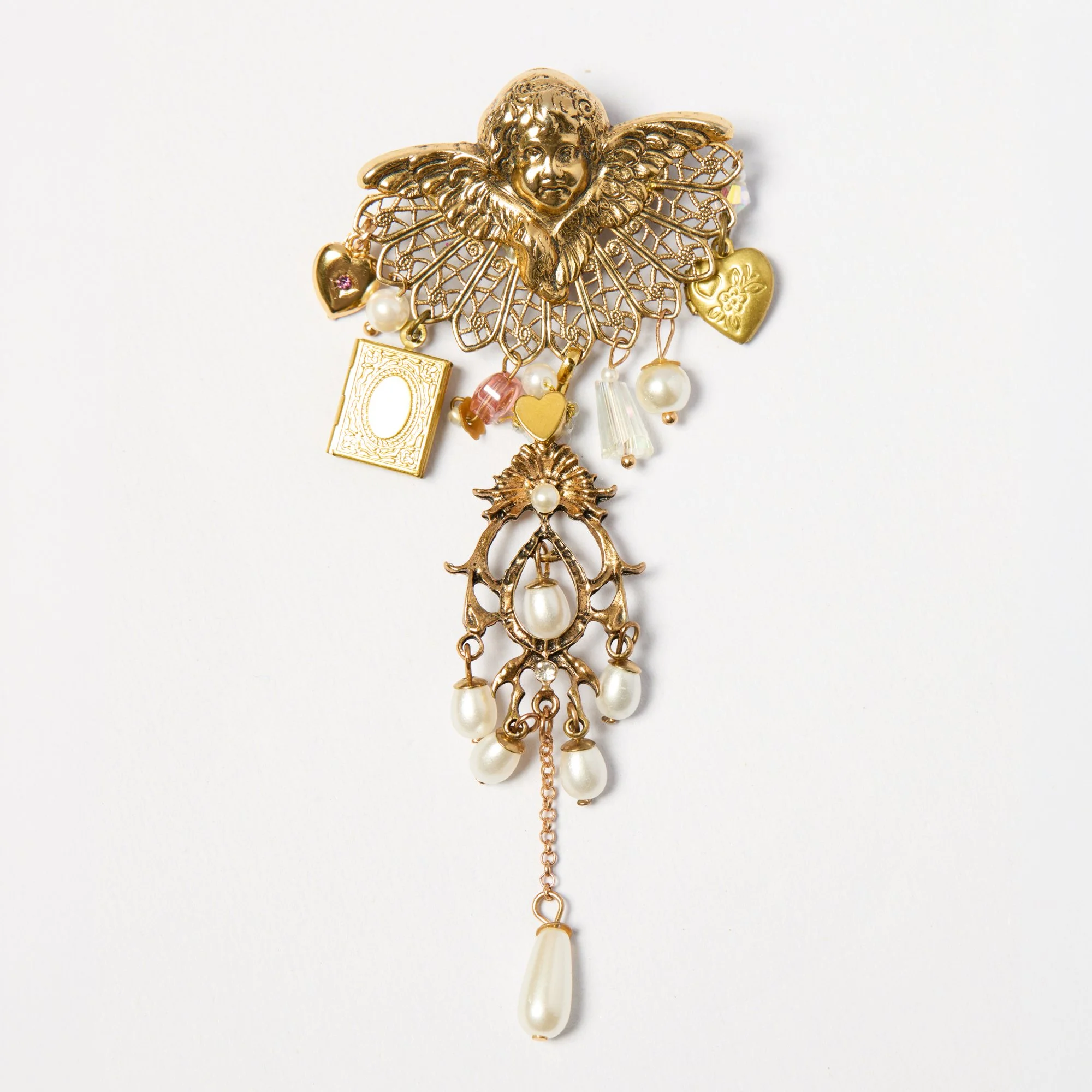 Gold decorative brooch with a cherub face, lace-like wings, and hanging charms including hearts, pearls, a book, and a rabbit.