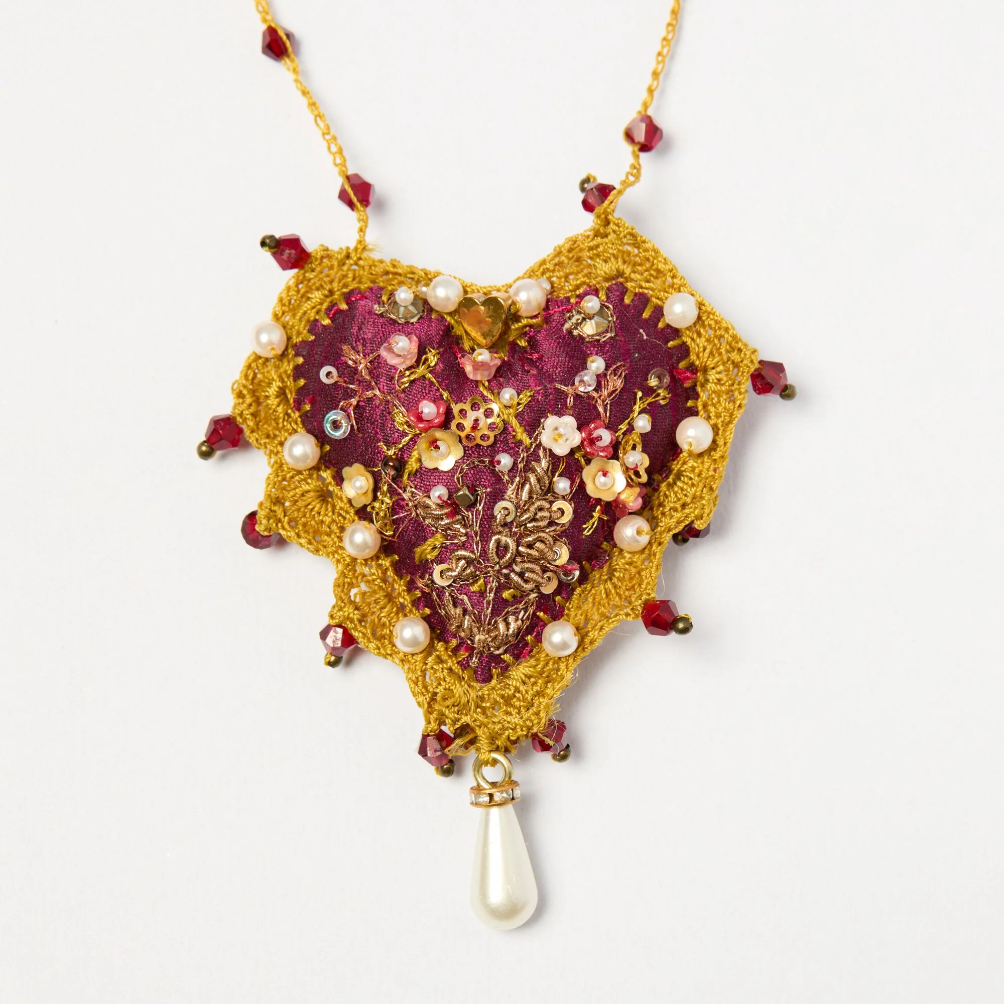 A decorative handmade necklace with a pink fabric base, surrounded by gold embroidery, pearls, and small flowers, attached to a golden chain with red beads and a large pearl drop hanging from it.