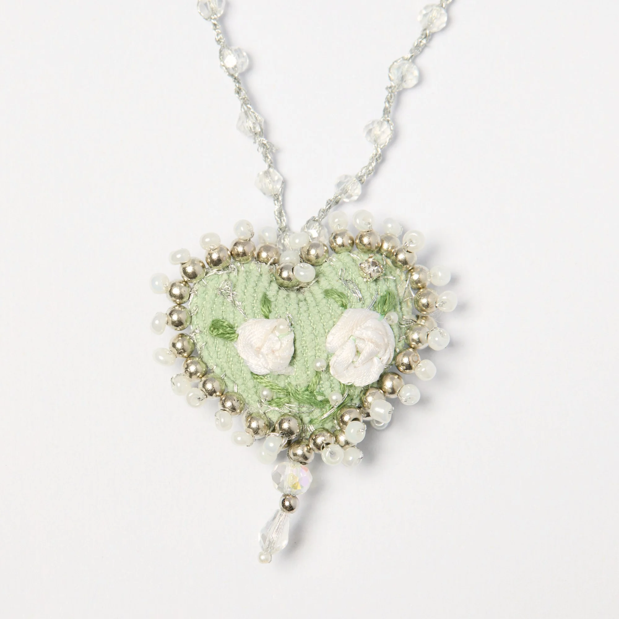 Close-up of an ornate piece of jewelry, possibly a necklace, with white, silver, and green beads, and a large clear crystal pendant hanging at the bottom, set against a white background.