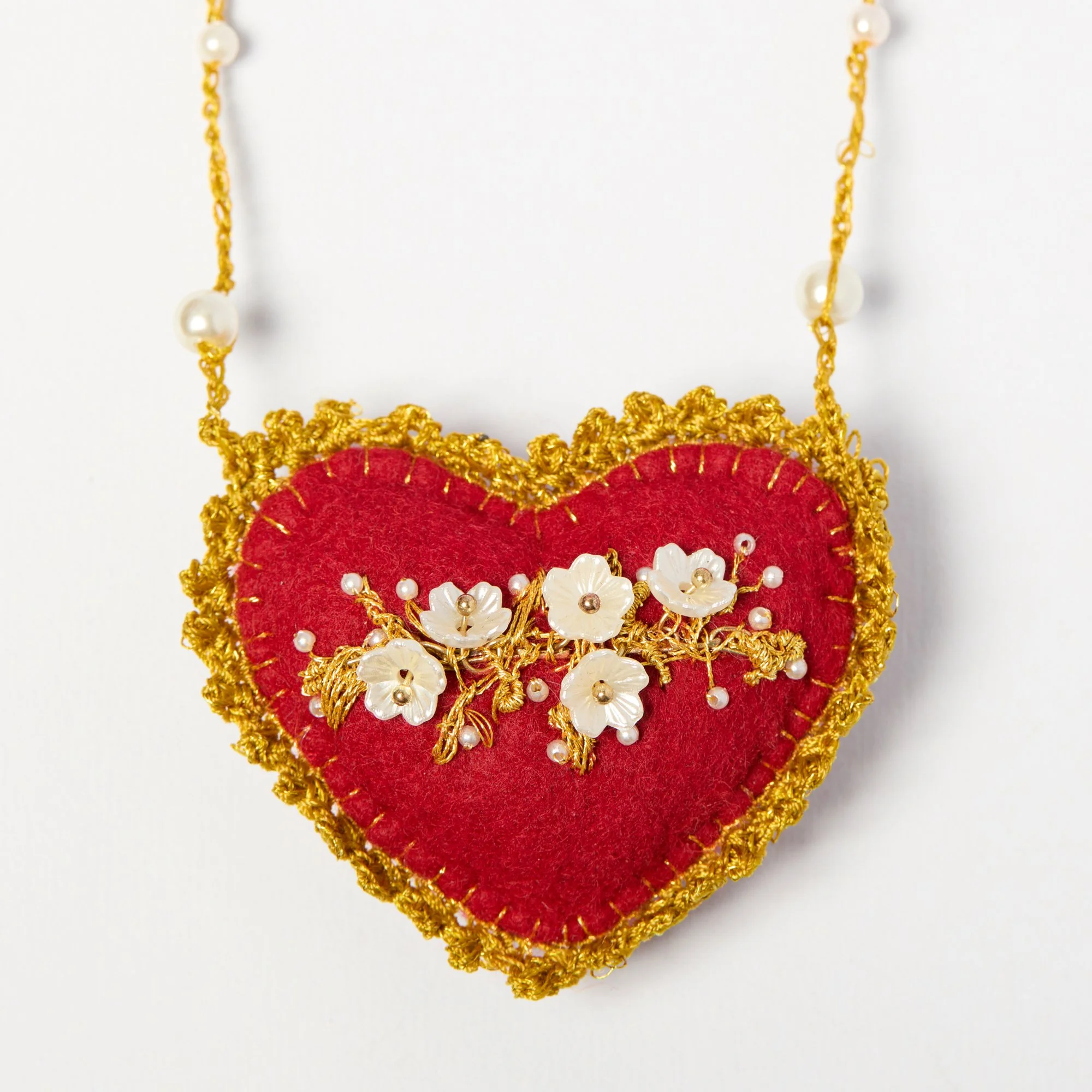 Close-up of a decorative hanging ornament with a red felt heart, gold embroidery, white flower-shaped beads, and pearl-white accents, against a white background.