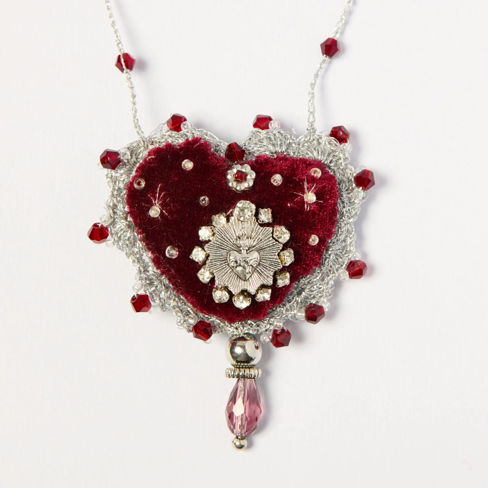 A decorative necklace featuring a heart-shaped velvet piece adorned with pearls, crystals, and red beads, with a pink crystal pendant hanging at the bottom.