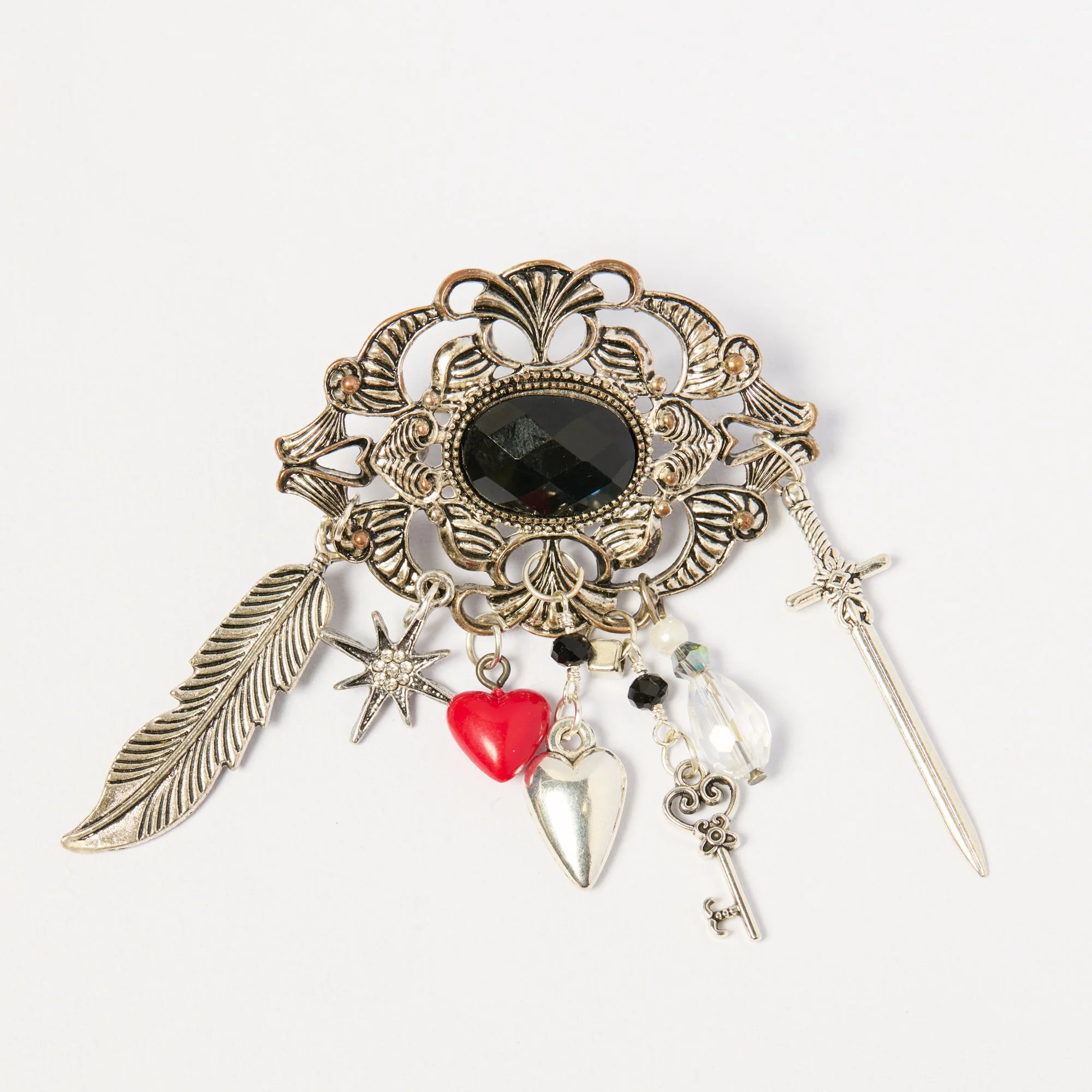 Close-up of a vintage brooch with silver feathers, a black gemstone, and dangling charms including a red heart, star, and key against a white background.