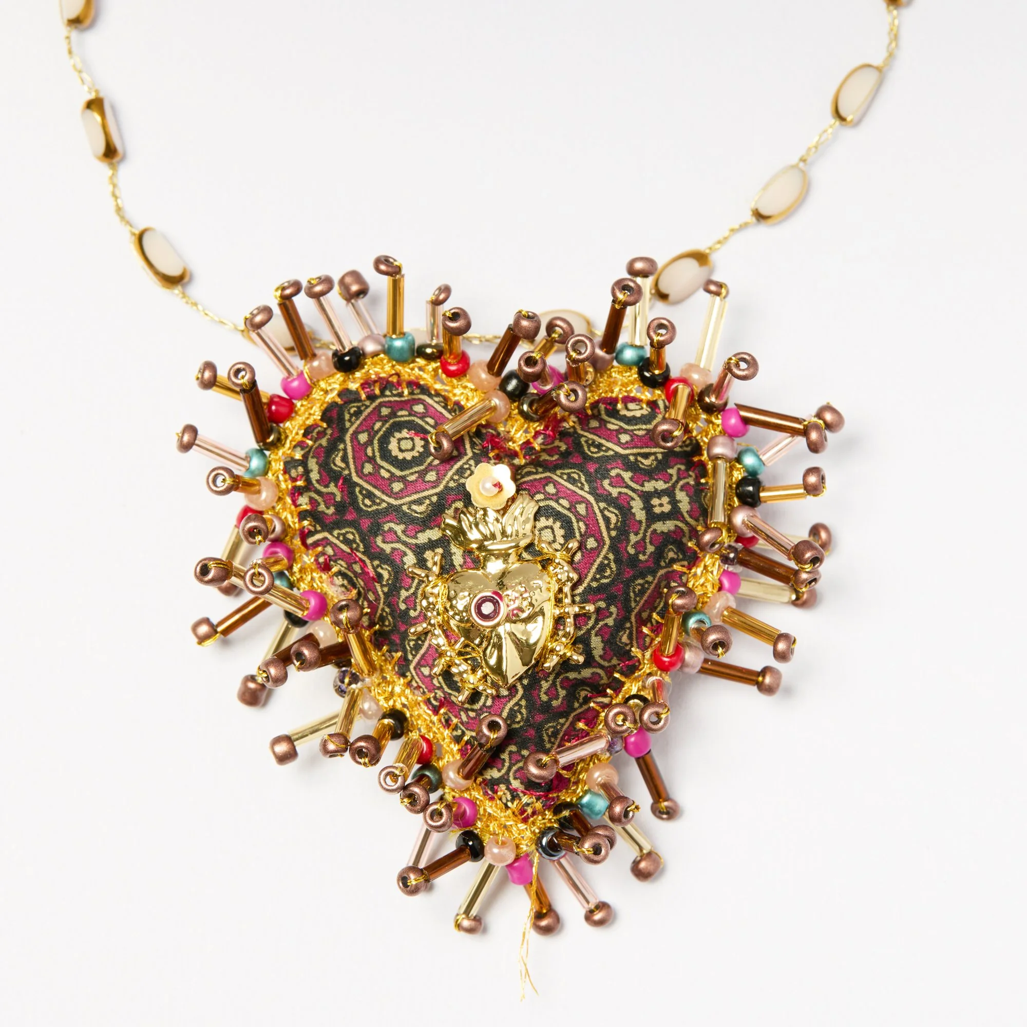 Close-up of an ornate decorative jewelry piece with gold, pink, and multicolored beads arranged in an intricate pattern, set against a white background.