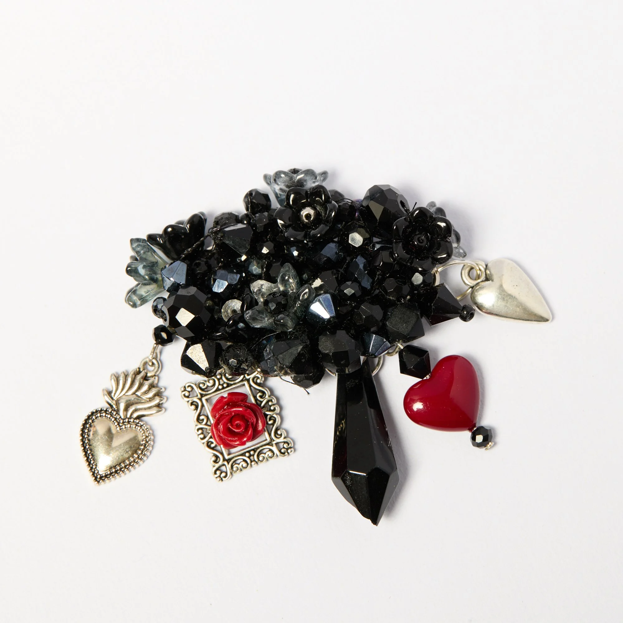 Close-up of a brooch, including black beads, silver charms, a red rose, and a large black pendant, arranged on a white background.