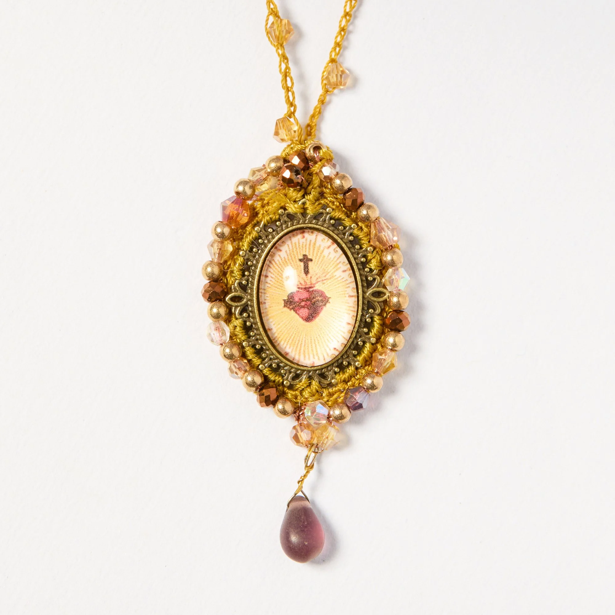 Close-up of a religious pendant necklace with a pink teardrop-shaped bead and gold-colored beads, adorned with intricate yellow threadwork.