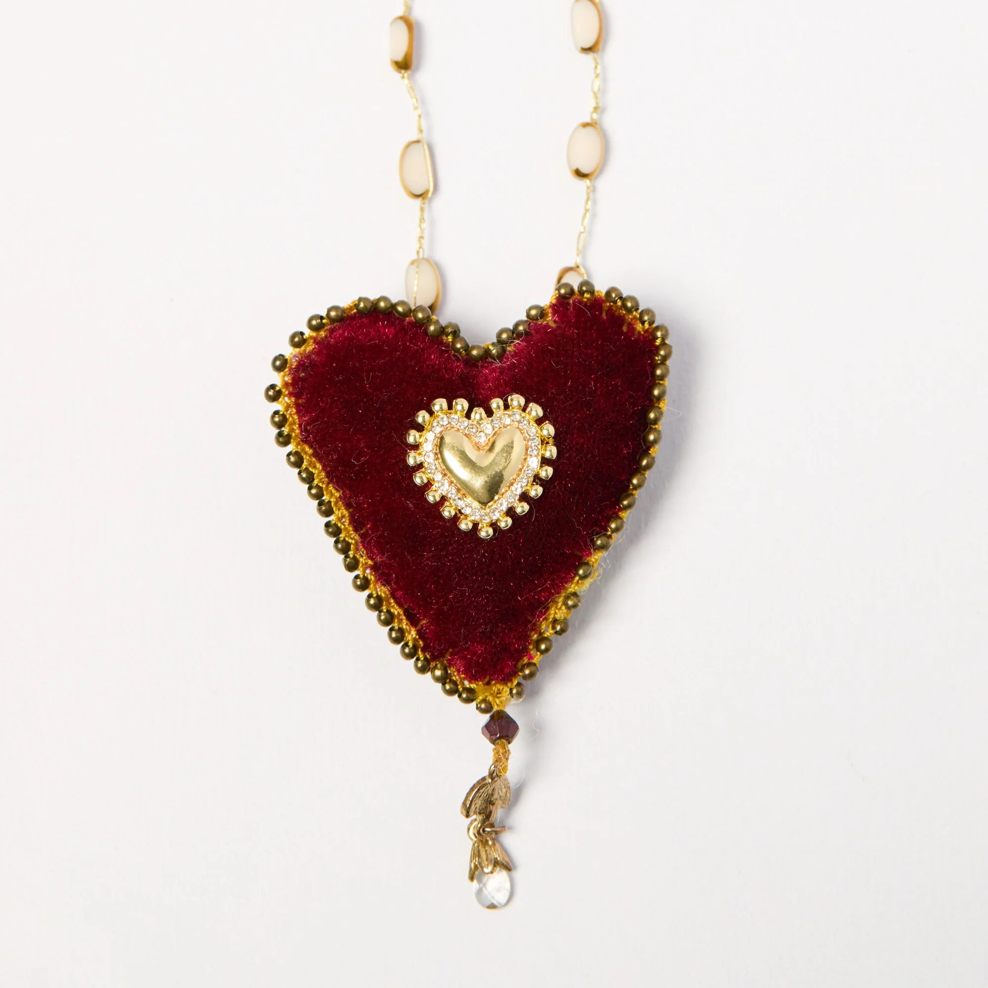 Gold chain necklace with a large velvet heart pendant decorated with gold beads and a heart-shaped design, hanging against a white background.