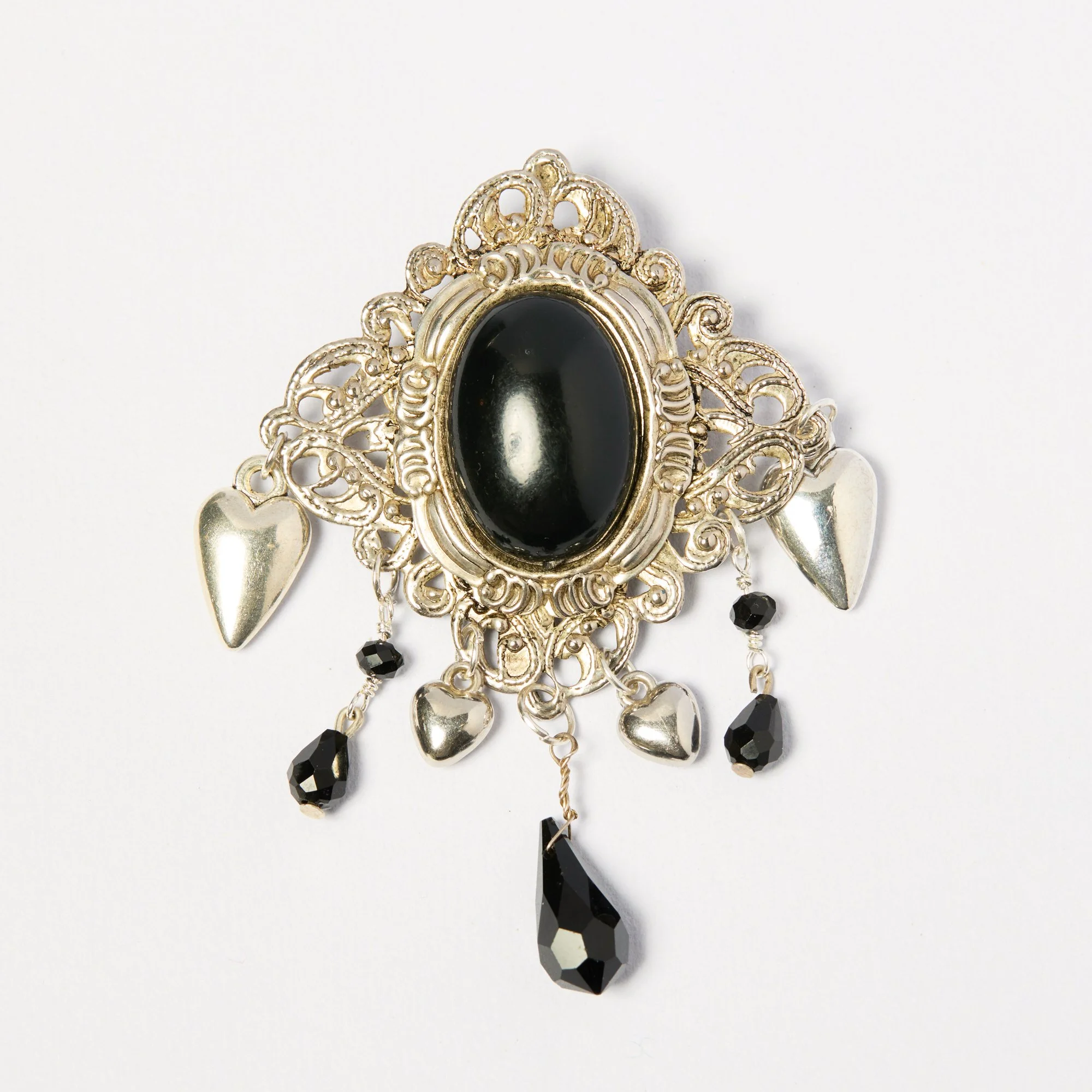 A decorative pendant with intricate silver filigree, featuring a black oval center stone, black faceted beads hanging at the bottom, and two silver heart-shaped charms on each side.