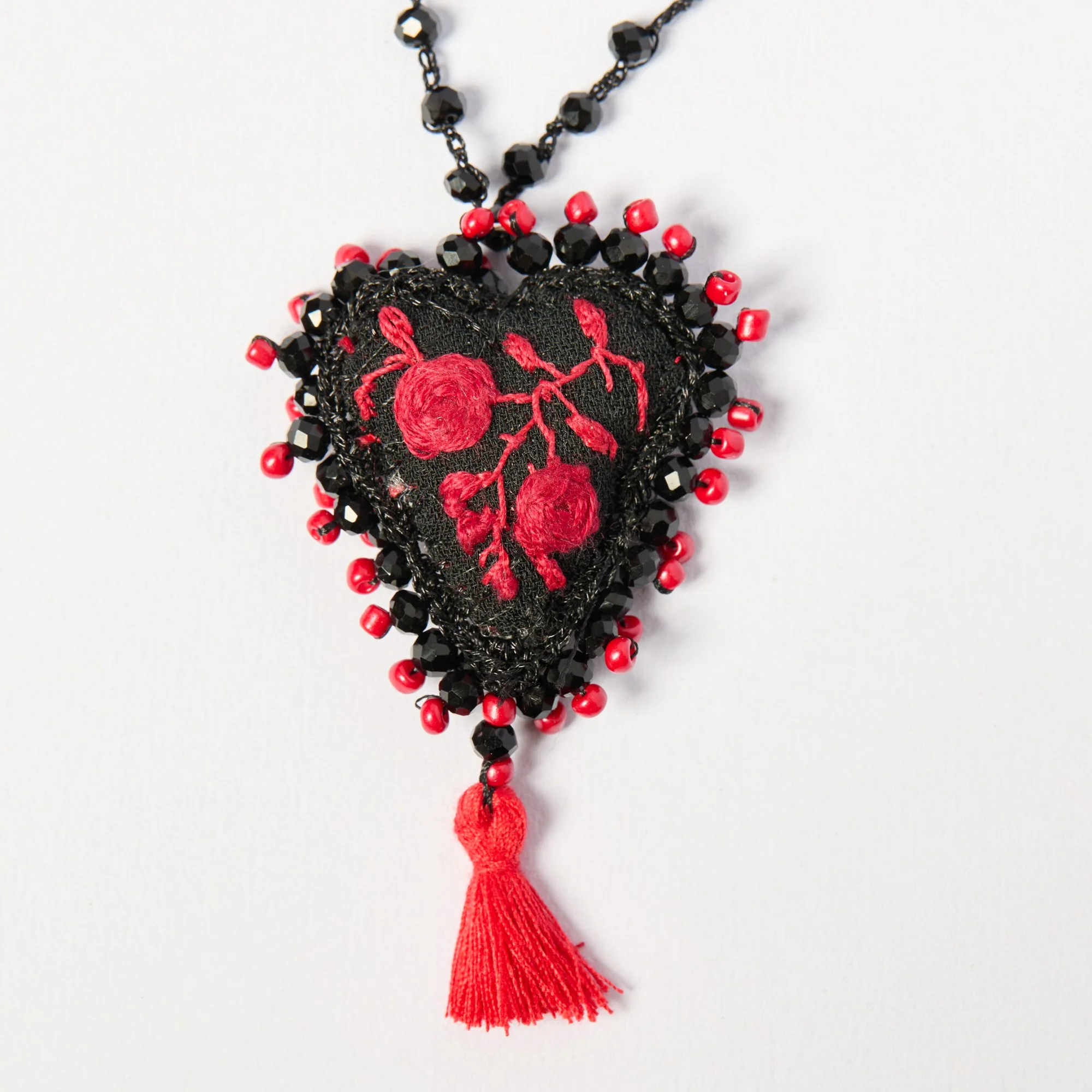 A handcrafted necklace with black beads, red beads, and embroidery of a red cherry branch on a black fabric heart, adorned with a red tassel.