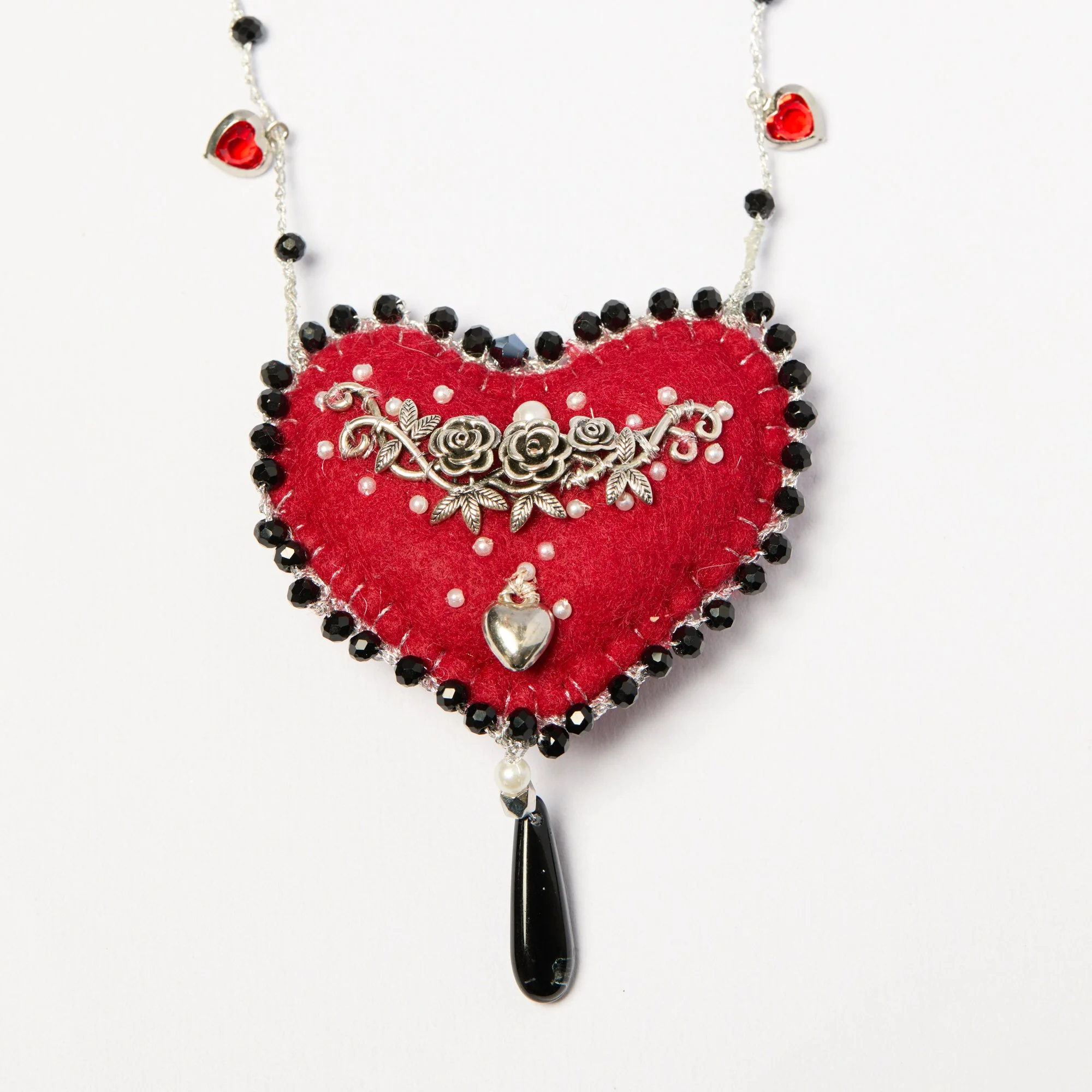 A necklace with a flower-shaped pendant, black beads, two red heart-shaped elements, and a silver decorative flower charm on a red felt heart background.