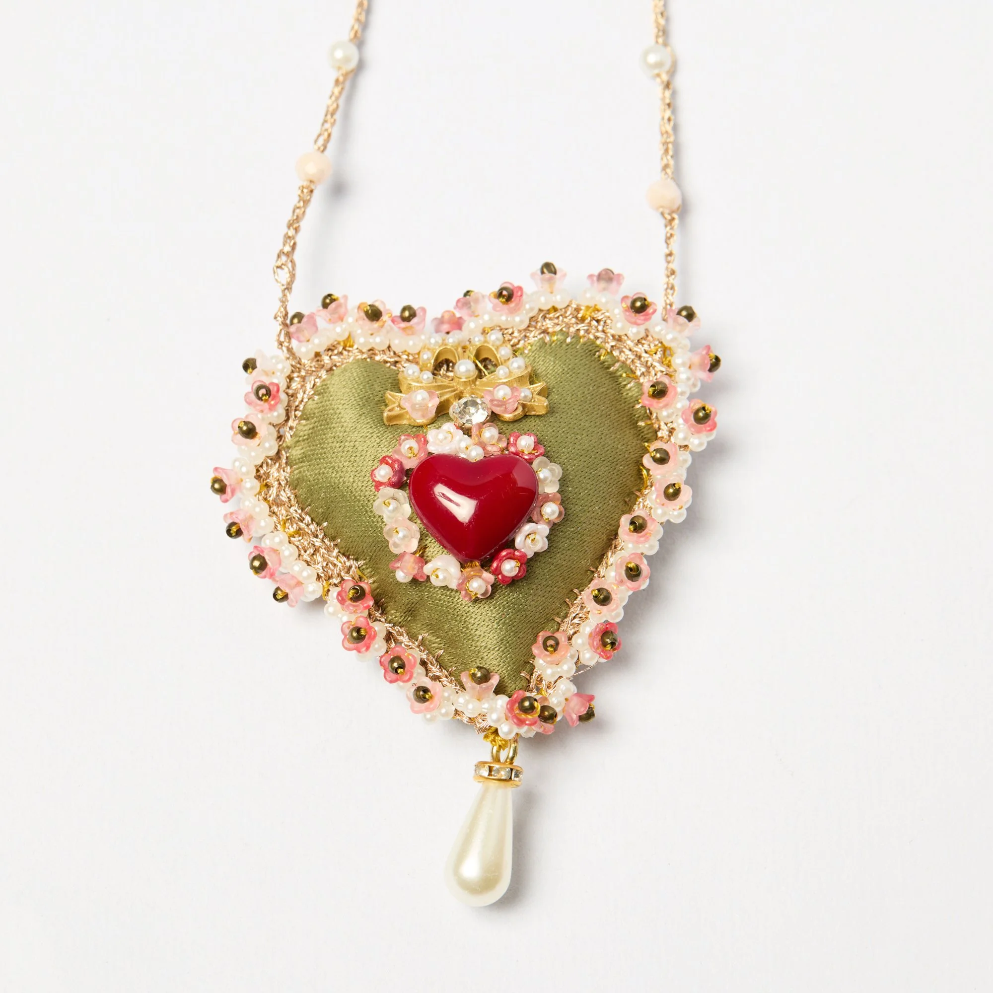 Close-up of a decorative necklace with a heart-shaped pendant, pearls, pink flowers, and gold accents on a plain white background.