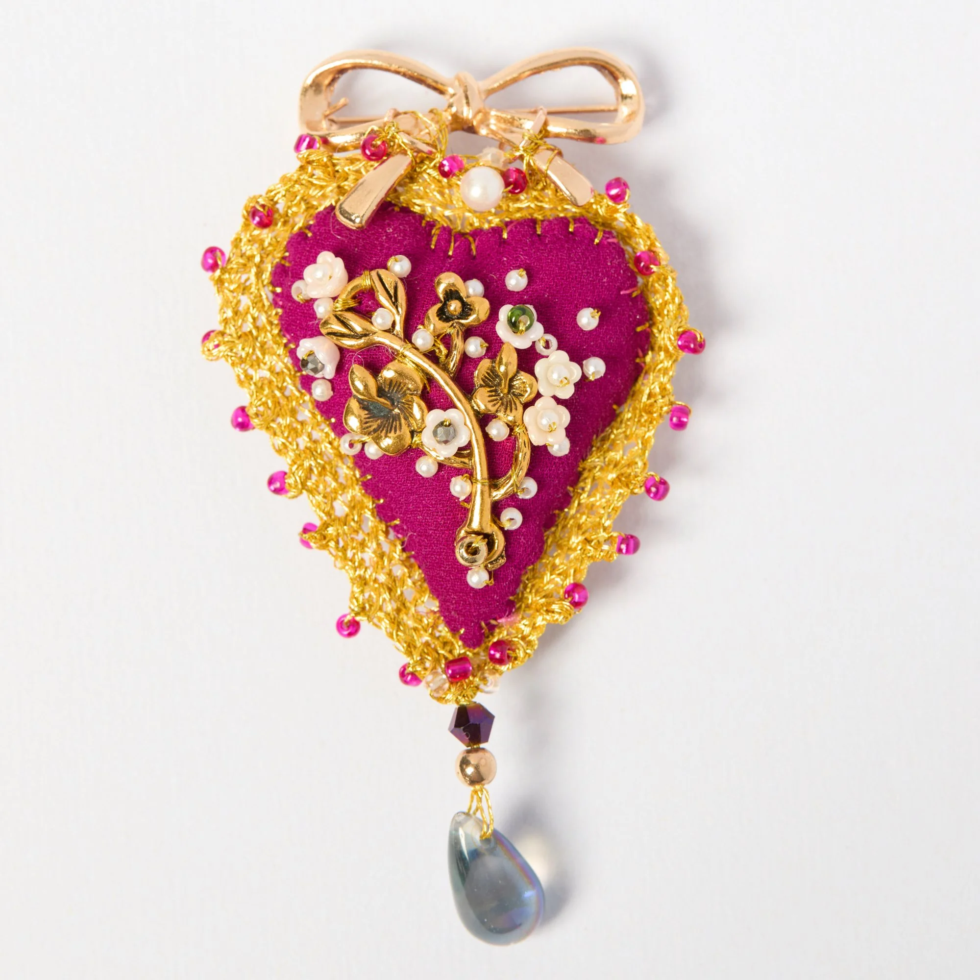 Decorative brooch with pink fabric, gold accents, pearls, pink beads, and a teardrop-shaped gem hanging from the bottom.