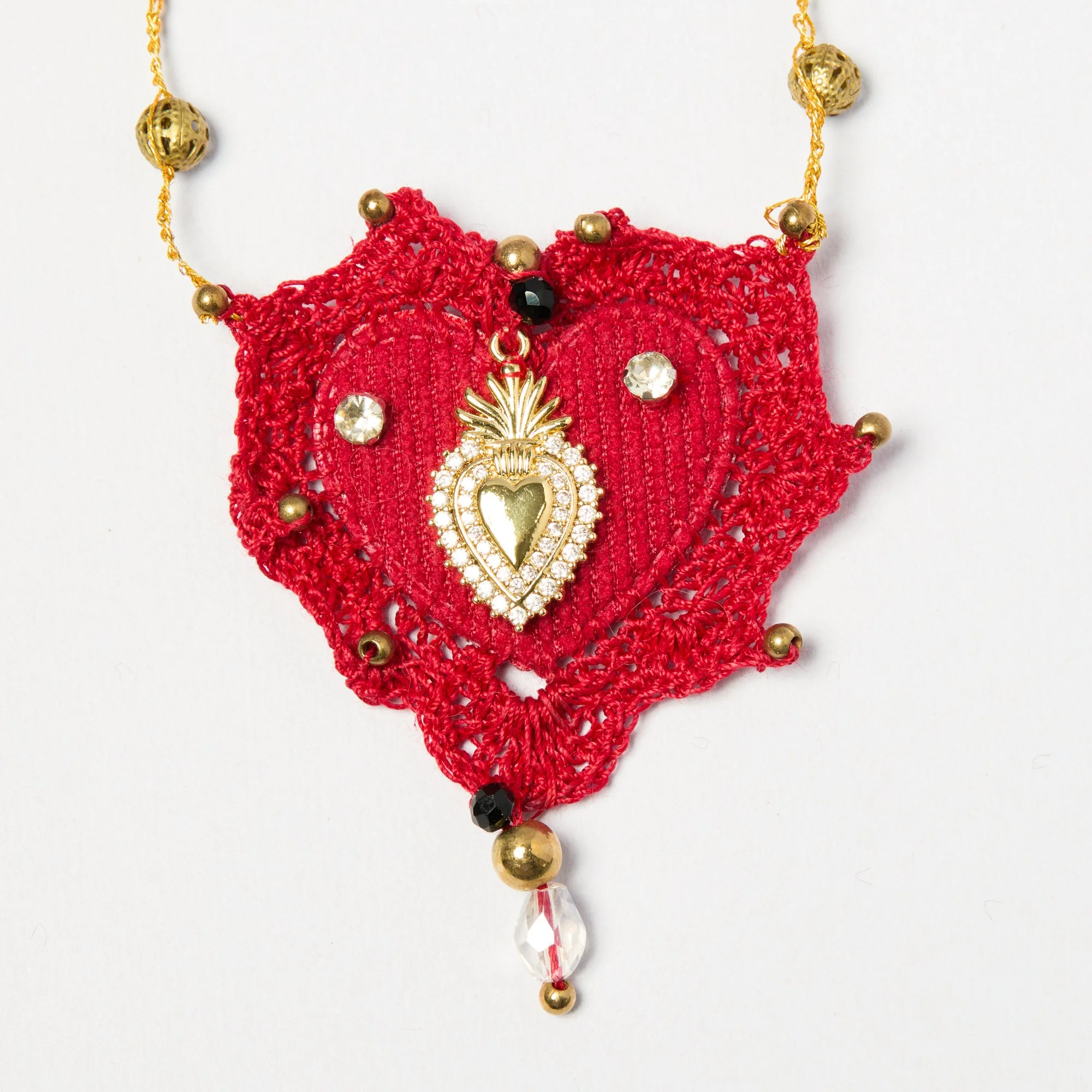 Close-up of a decorative necklace with red crocheted fabric, gold beads, black beads, a star-shaped pendant with rhinestones, and a clear teardrop-shaped pendant on a white background.