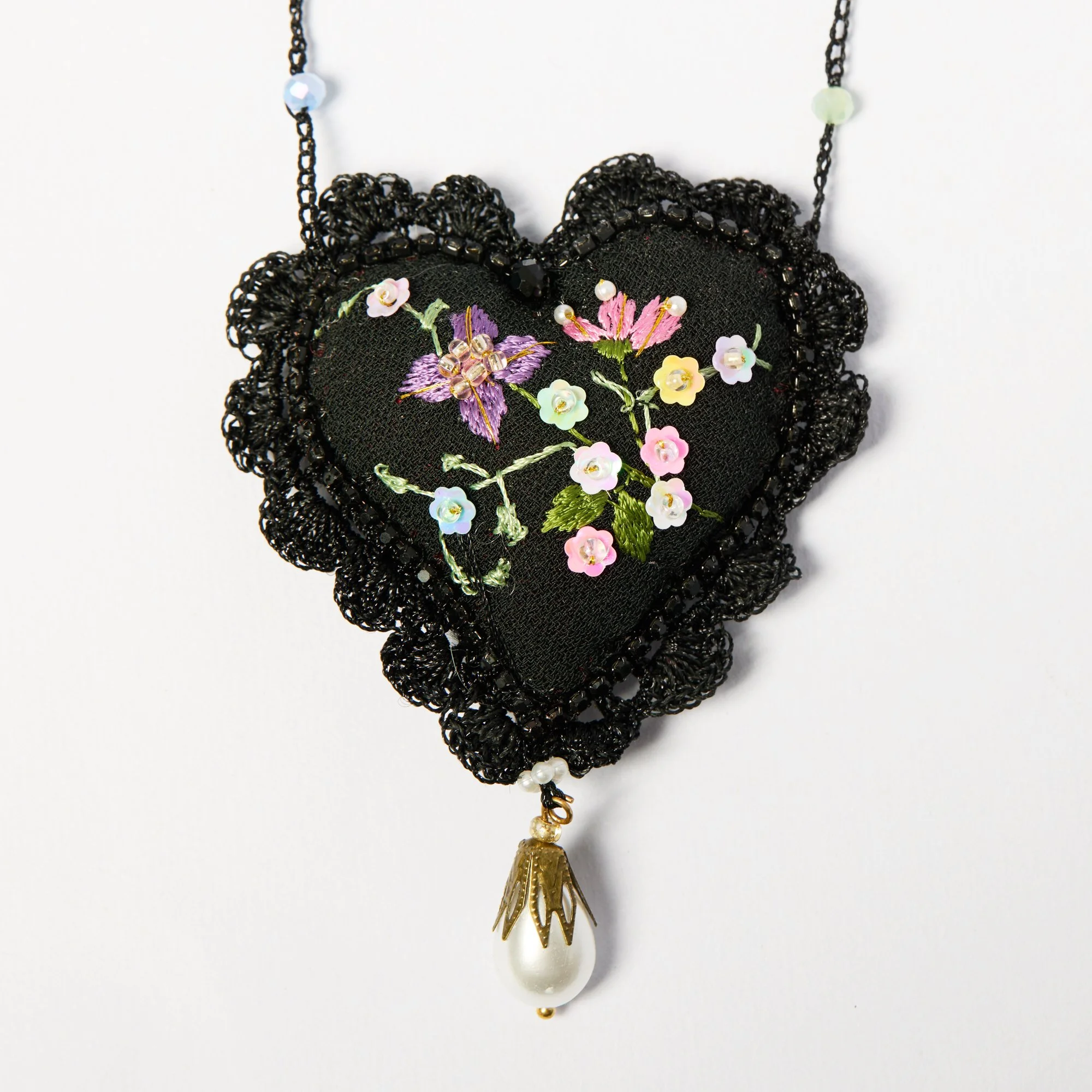 Close-up of a black fabric heart-shaped handmade necklace with floral embroidery and beaded decoration, hanging from a chain with pastel beads and a pearl pendant.