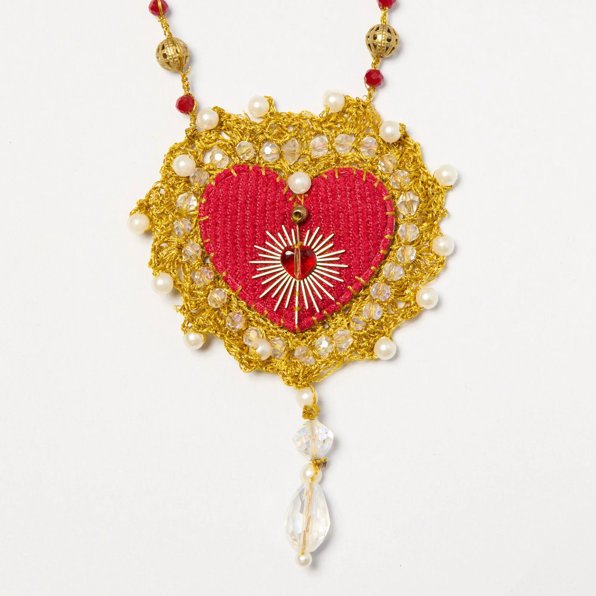 Close-up of ornate necklace with gold, pearls, red beads, and crystals arranged in a floral pattern, with hanging teardrop-shaped crystals and a red heart-shaped pendant.