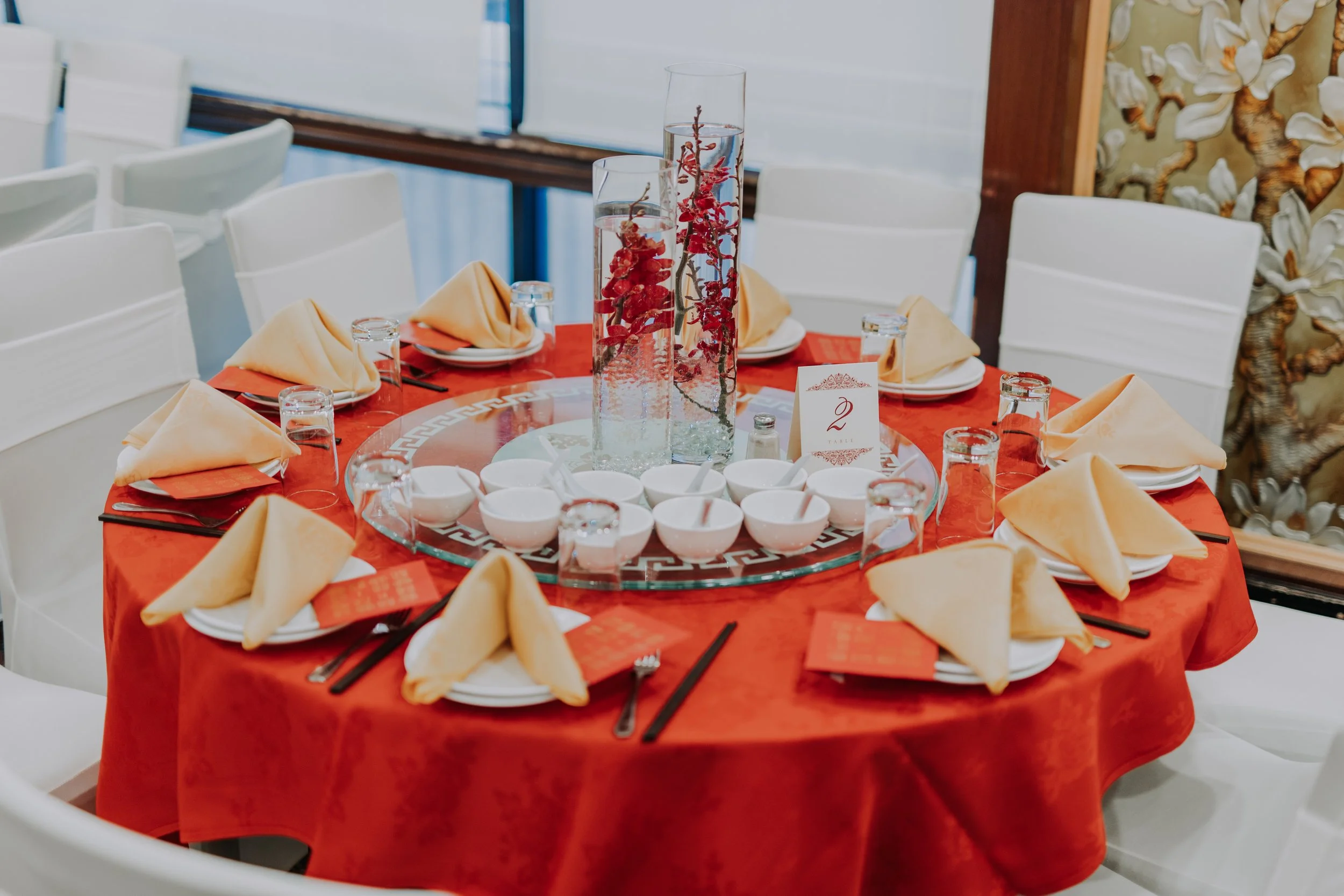 A round dining table set with yellow folded napkins, small bowls, glasses of water, and chopsticks on a red tablecloth in a restaurant with white chairs and a decorative wall panel.