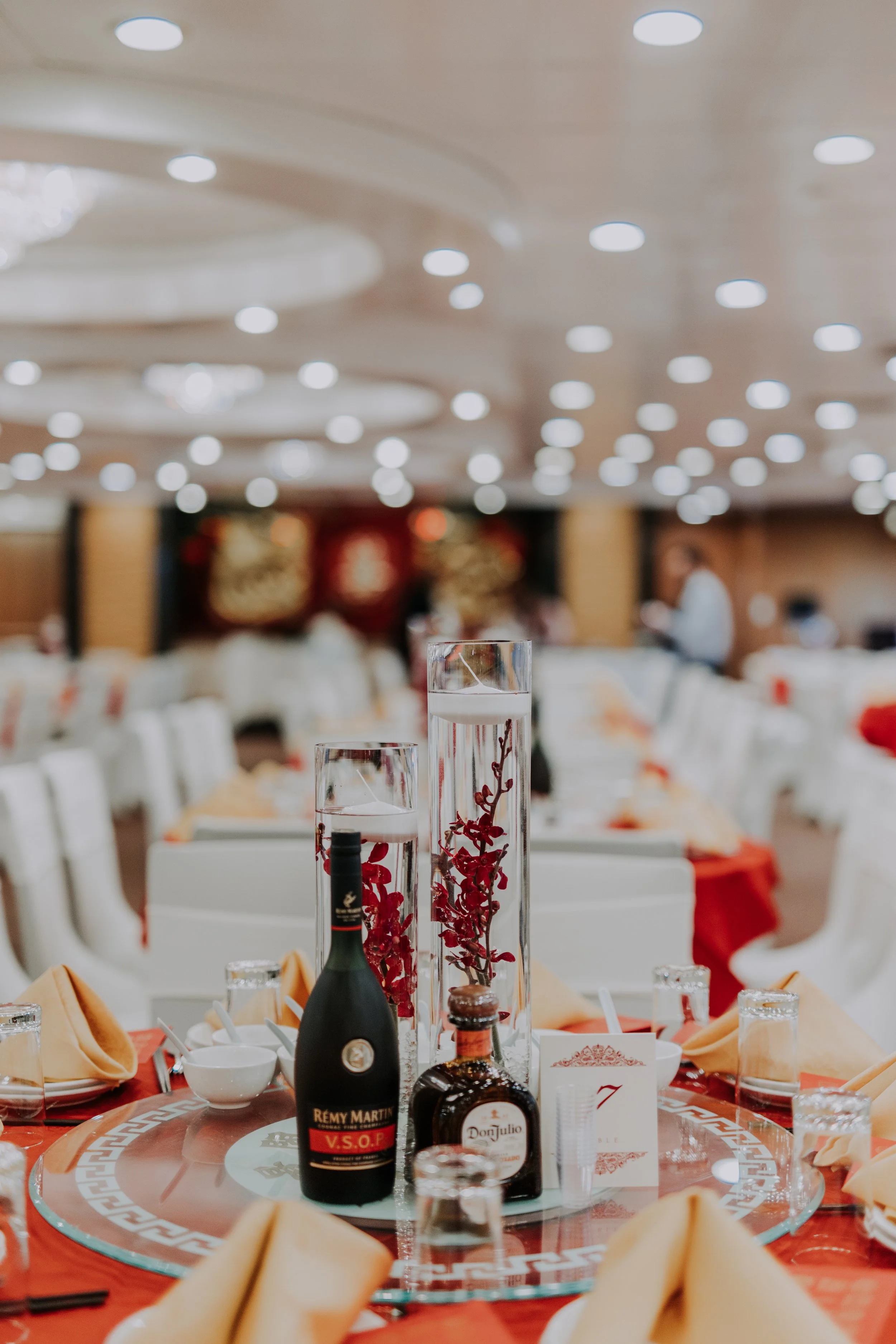 A banquet table with bottles of alcohol, decorative centerpieces, napkins, and place settings in a banquet hall with bright ceiling lights.