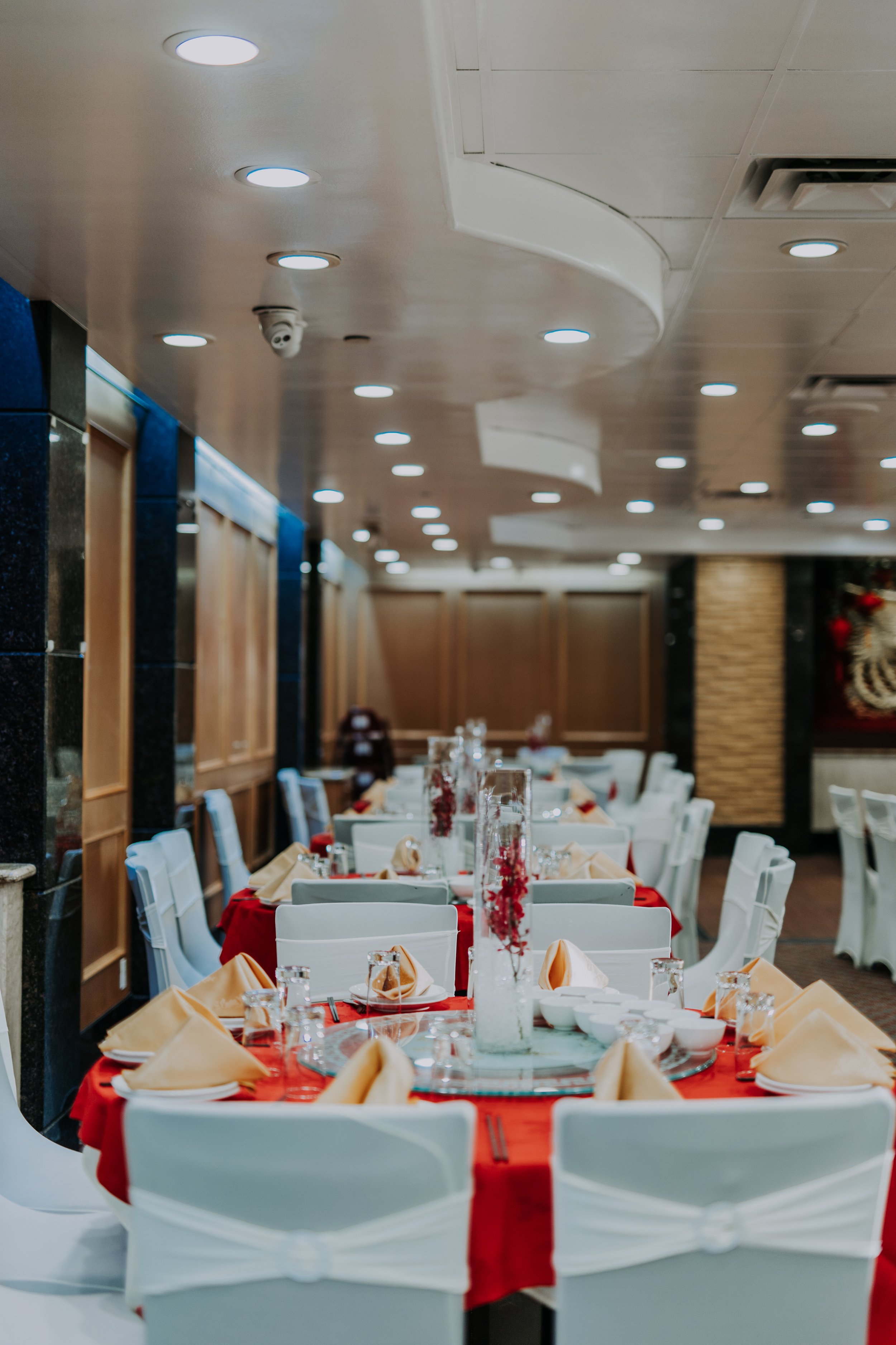 Restaurant or banquet hall decorated for a special occasion with tables set with beige napkins, glassware, and centerpieces featuring red flowers.