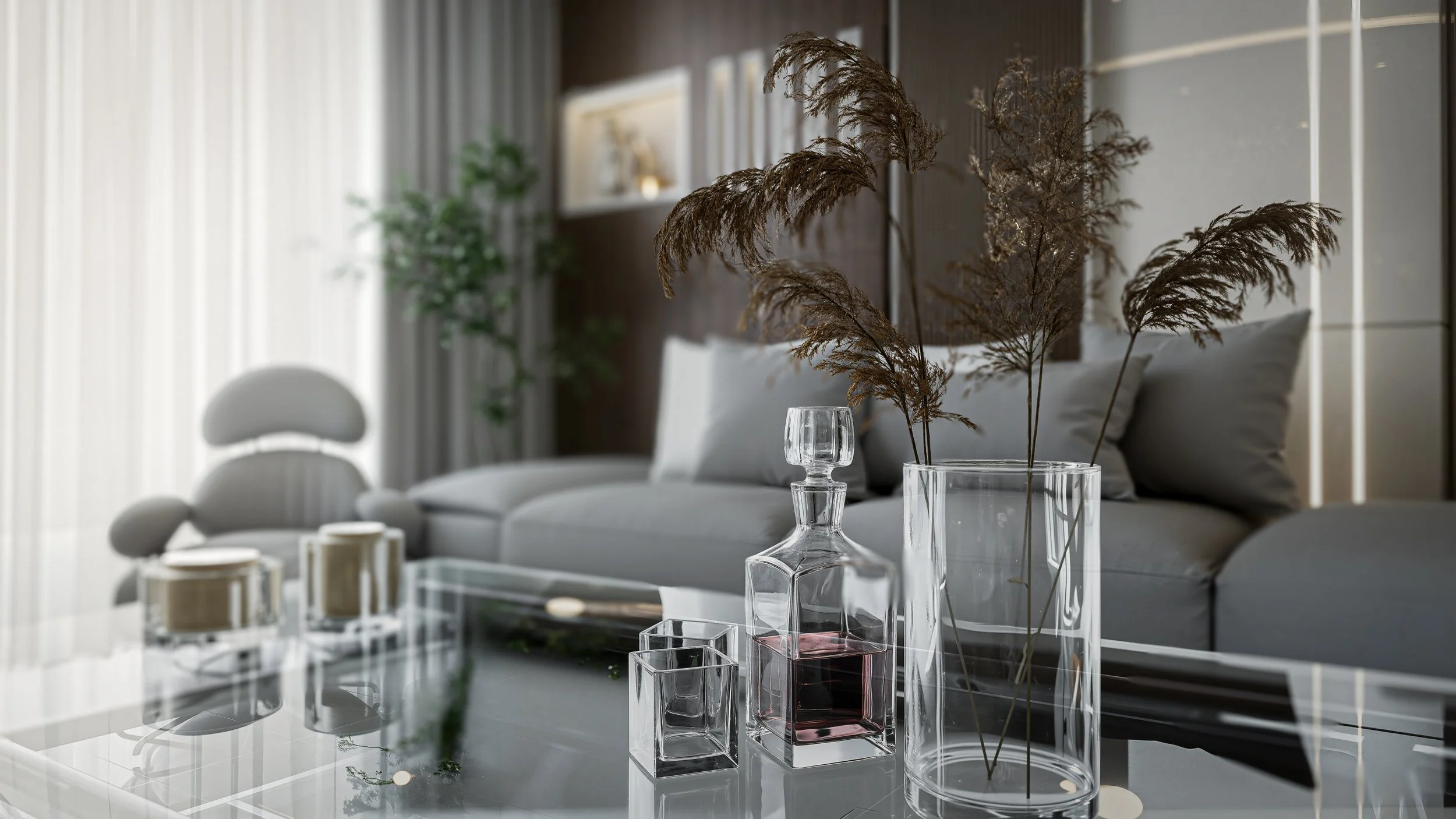 Interior of a modern living room with a glass table holding decorative vases with dried plants, a gray sofa with cushions, and a blurred background of a window with curtains and plants.