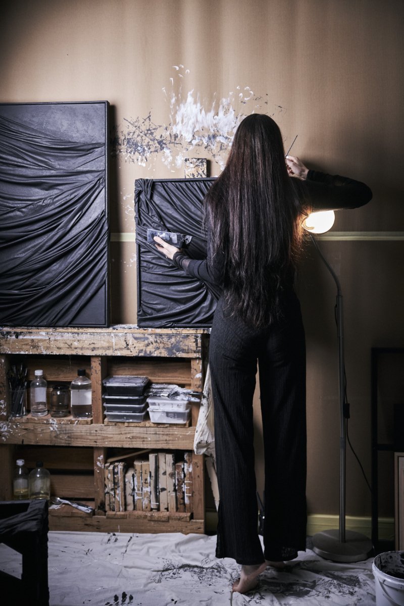 Artist with long dark hair painting a large abstract black-and-white artwork on an easel in a studio, surrounded by art supplies and paint splatters.