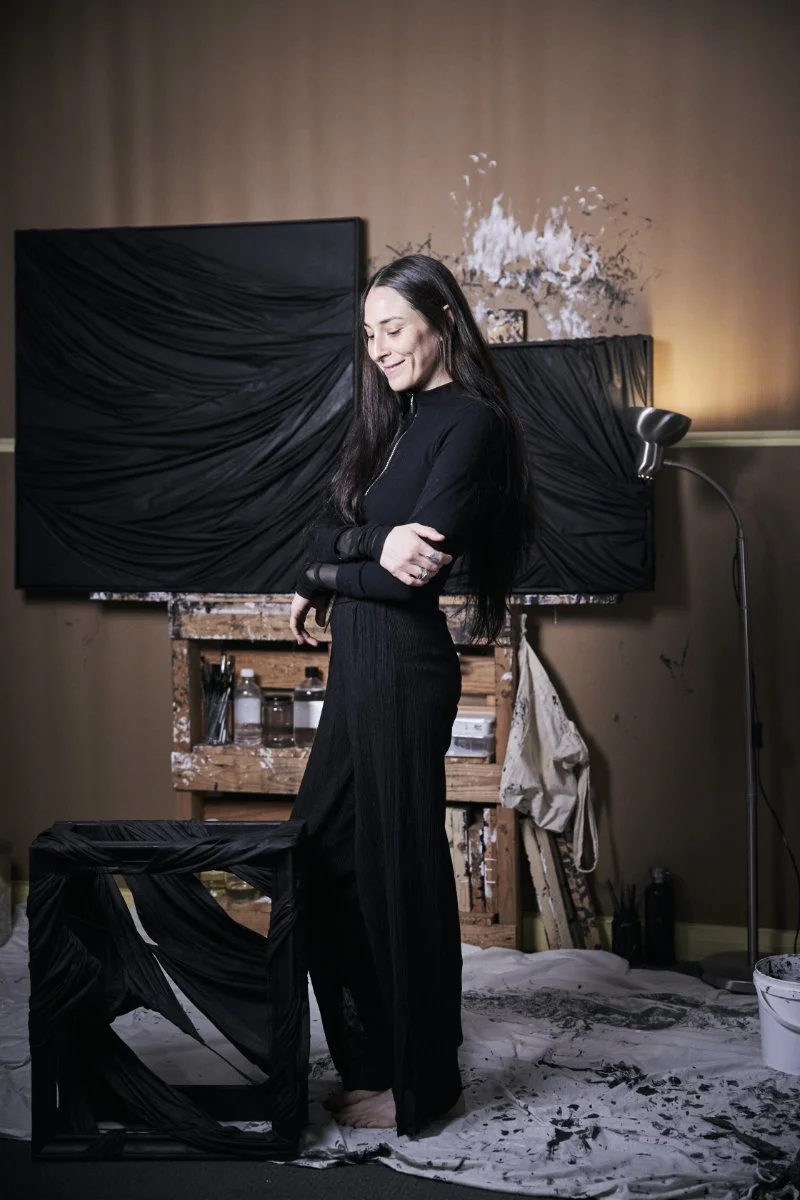 A woman with long dark hair, wearing a black long-sleeve dress, stands in an art studio. She is smiling with her eyes closed and her arms crossed. Behind her is a large canvas with black draped fabric, and in front of her is a black chair covered with similar fabric. The studio has a paint-splattered canvas, bottles of paint, and art supplies on a wooden easel and shelf. The floor is covered with a white tarp stained with black paint.