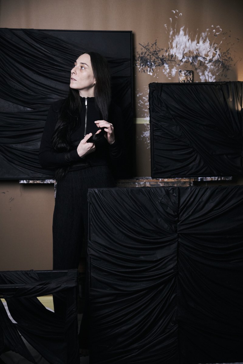 A woman with long black hair stands among black wrapped objects in an art studio, with a wall decorated with an abstract splash of white and black paint behind her.