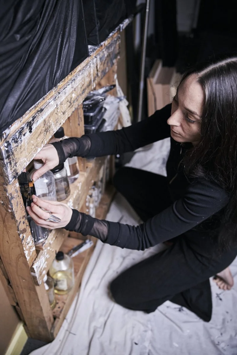Woman kneeling down and working on a wooden workbench with various jars and tools, partially covered in paint or plaster, in a workshop