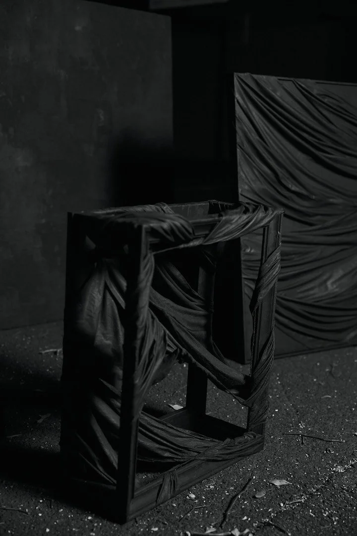 A black, fabric-covered empty display stand or frame set against a dark wall, with another wrapped object in the background, in a dimly lit room with scattered debris on the floor.