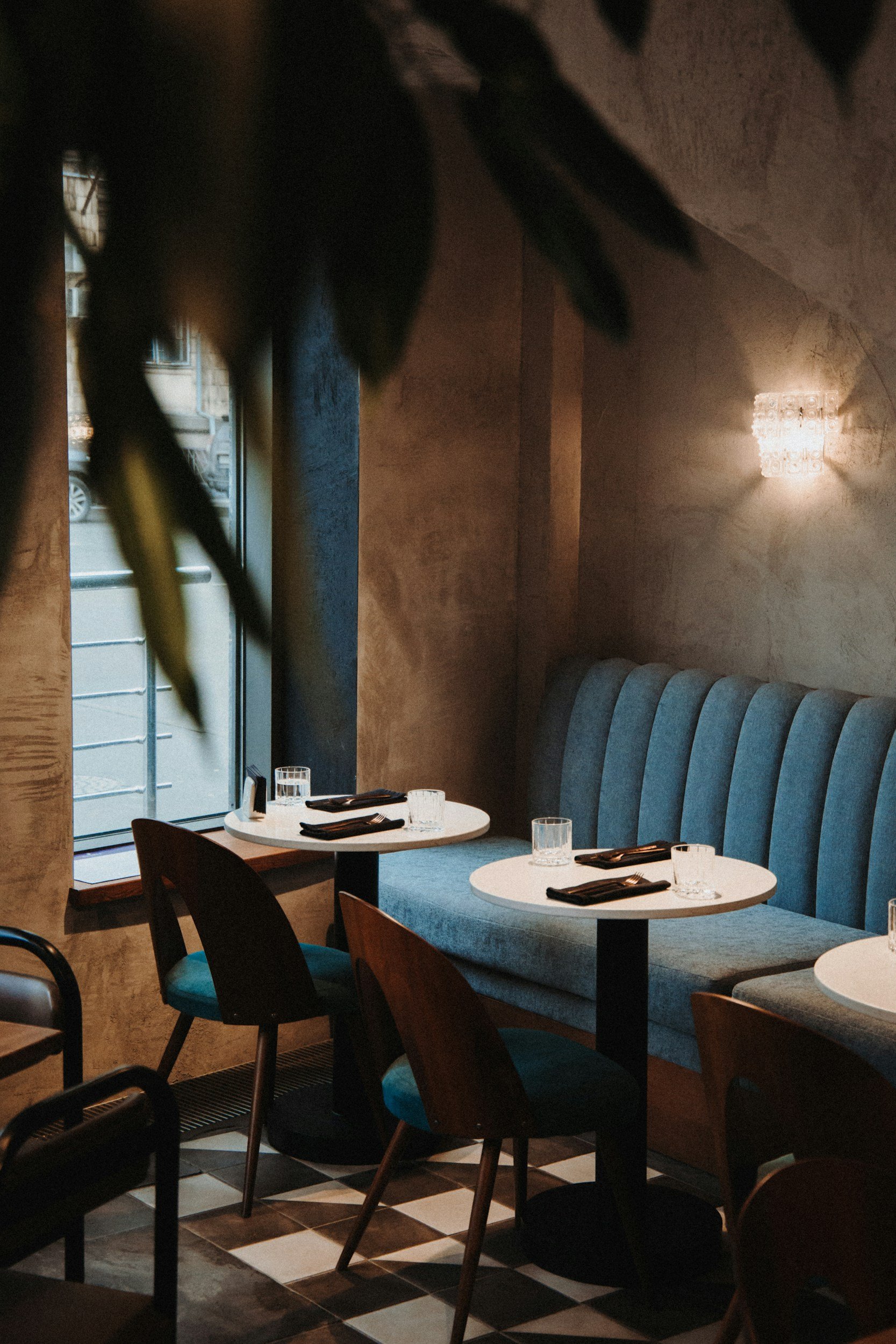 Interior of a restaurant with white round tables set with glasses and black napkins, black wooden chairs, blue cushioned bench seating along the wall, large window with city view, and a textured wall with modern light fixture