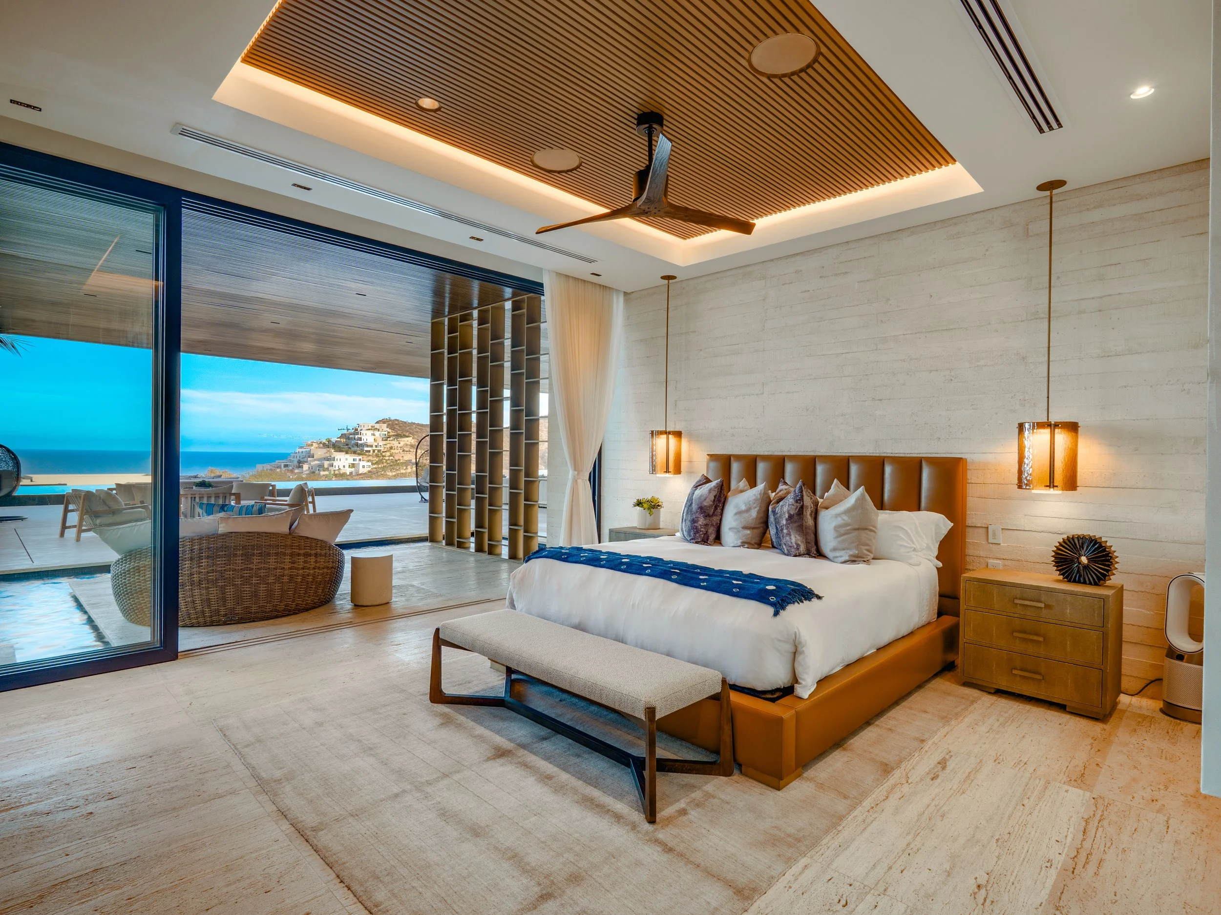 A modern bedroom with a large bed featuring white bedding and multiple pillows, a wooden side table, and a bench at the foot of the bed. Floor-to-ceiling glass doors open to a balcony with outdoor seating and a view of the ocean and hillside buildings. The ceiling has wooden slats with integrated lighting and a ceiling fan, and the room has neutral, earthy tones.