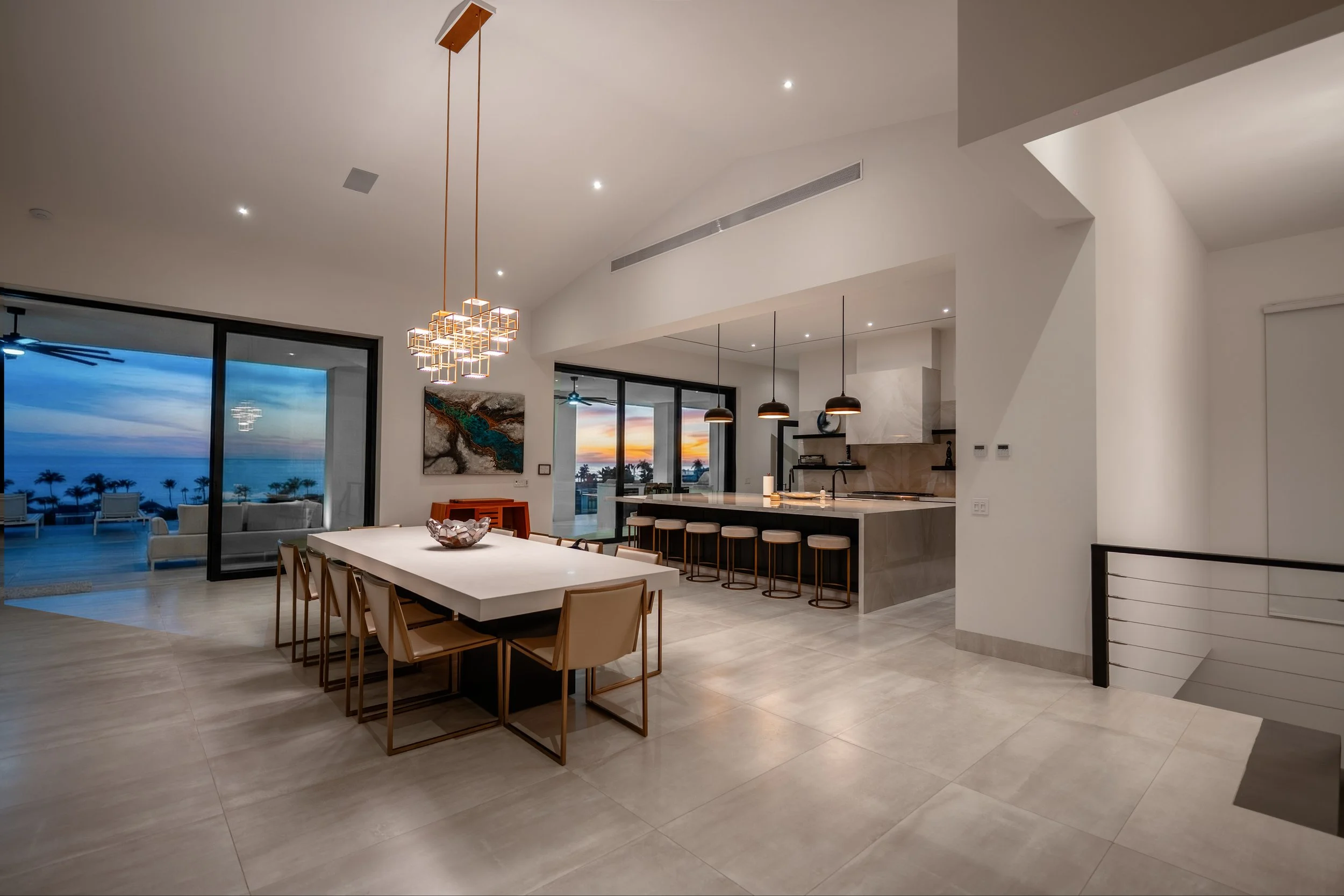 Modern open-concept dining and kitchen area with large windows showing a sunset and beachfront view, featuring a white dining table with beige chairs, geometric gold chandelier, and a minimalist kitchen with black pendant lights.