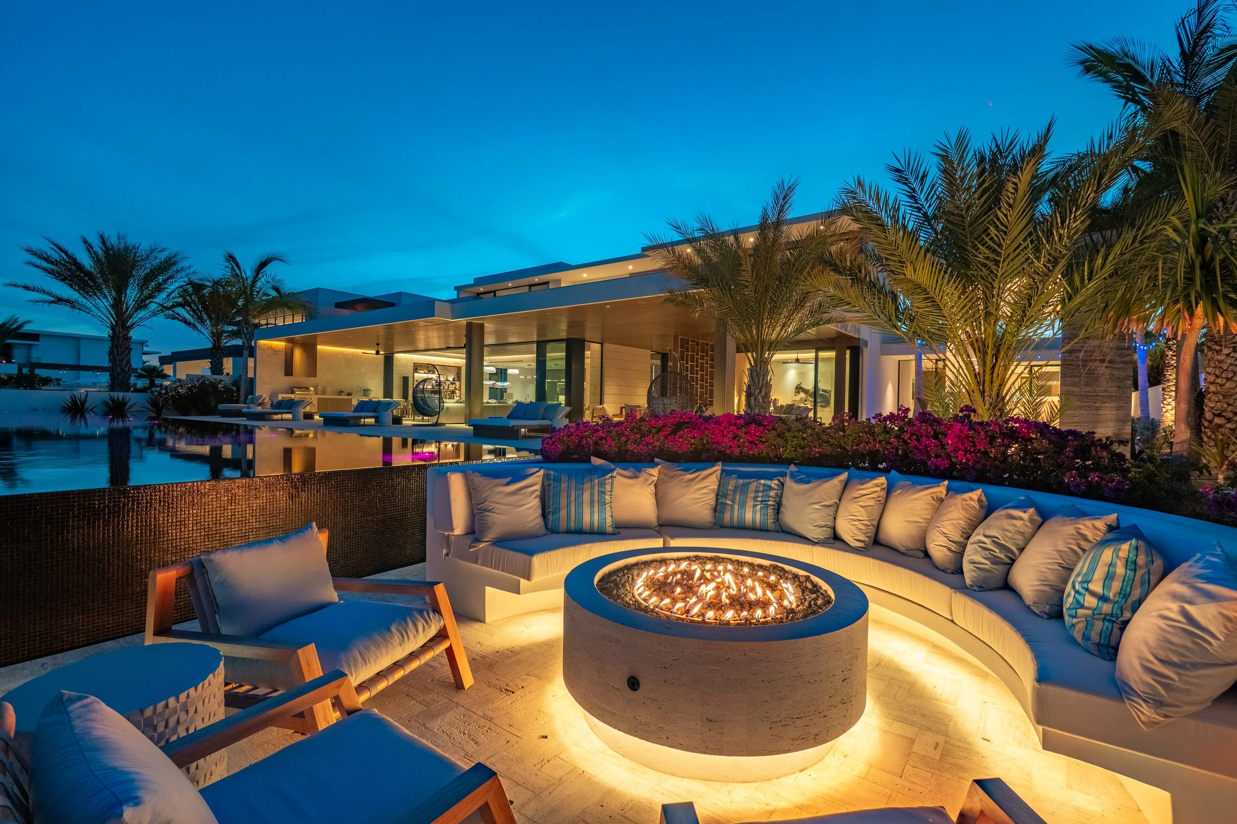 Luxury outdoor patio at dusk featuring a round fire pit, cushioned seating, palm trees, colorful flowers, and a modern house with large glass windows and warm lighting.