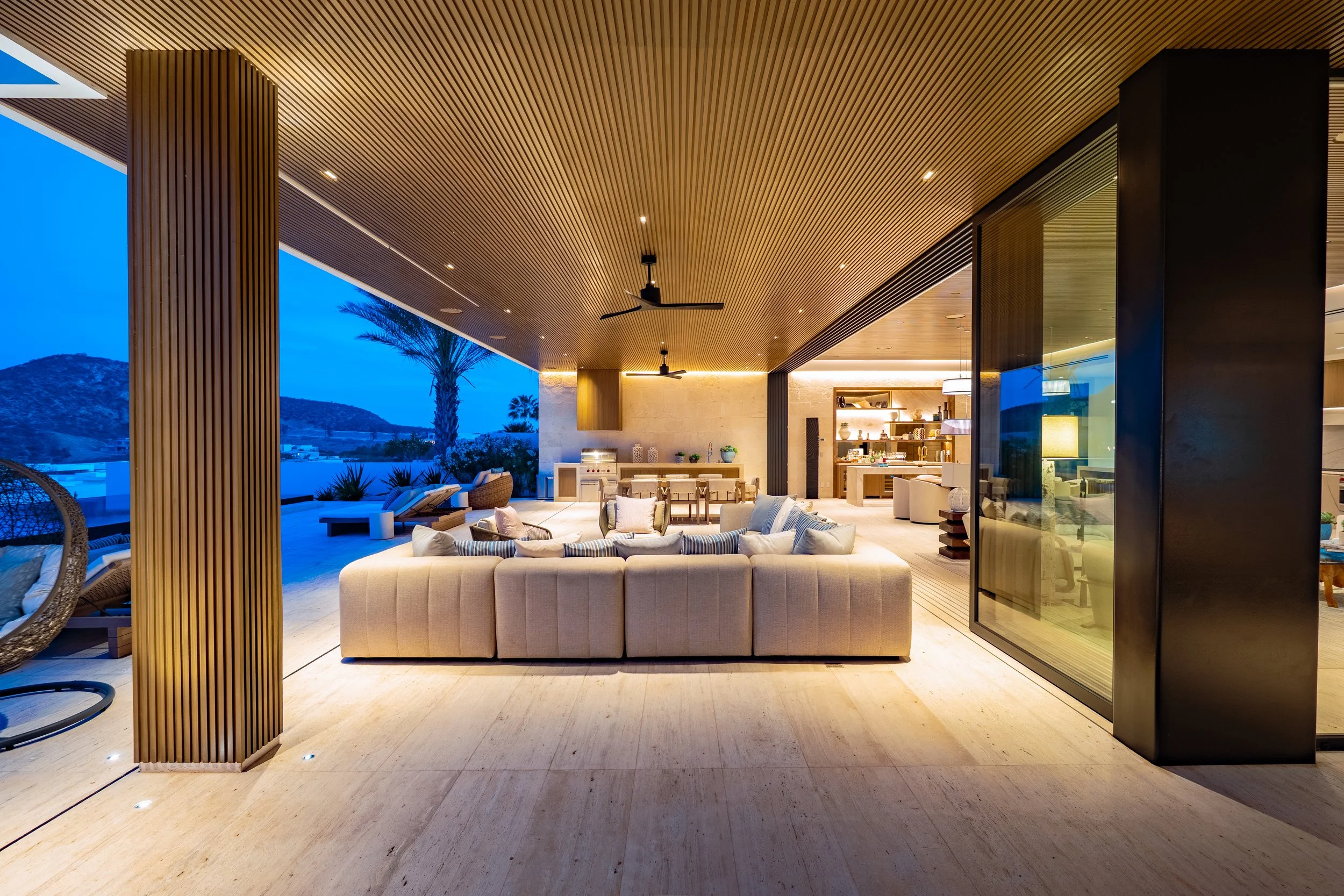 Modern outdoor living space with a beige sectional sofa, wooden ceiling, ceiling fans, and a view of a mountainous landscape at dusk.