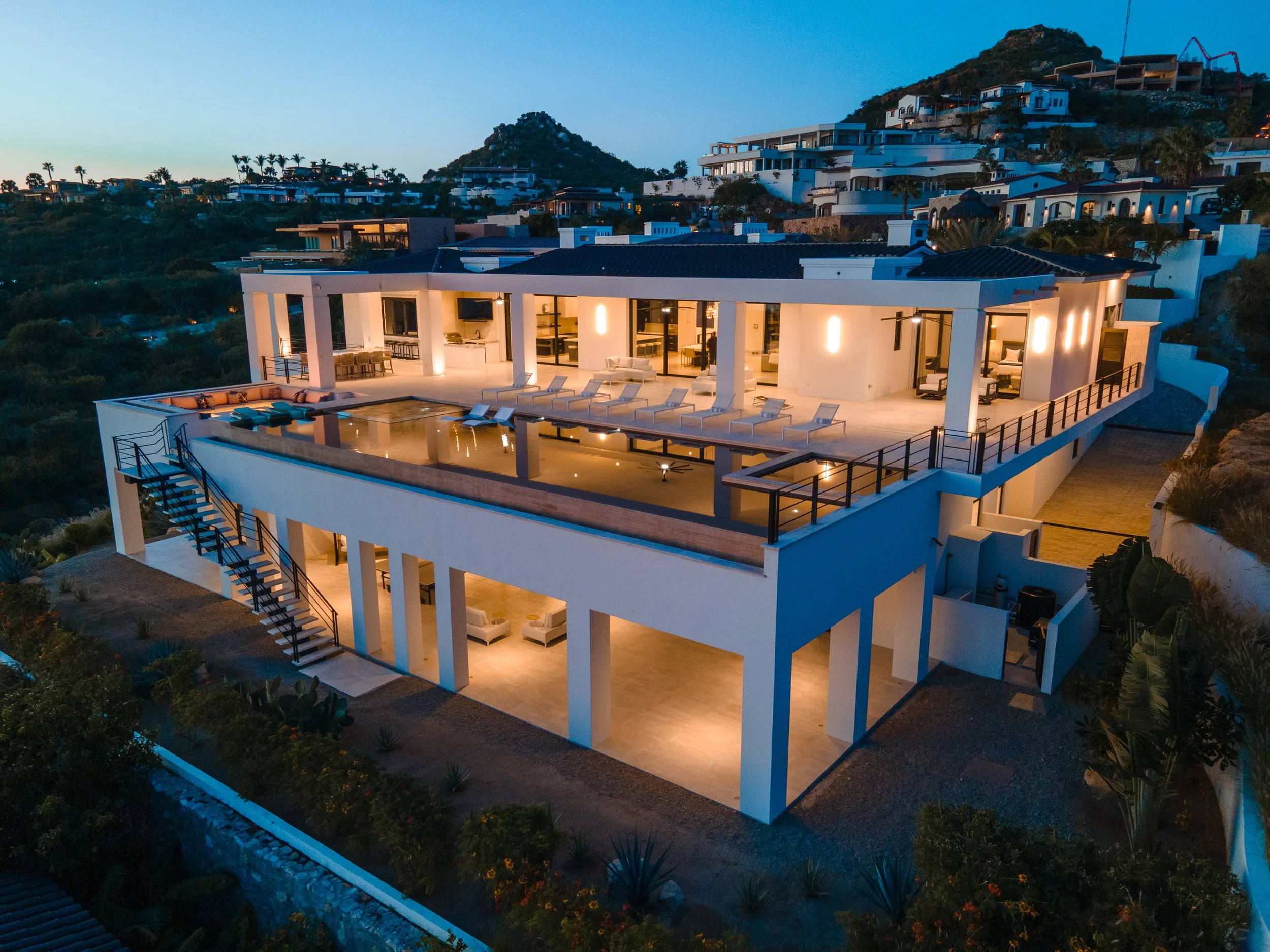 A modern house on a hillside at dusk with an illuminated outdoor patio, pool, and lounge chairs.