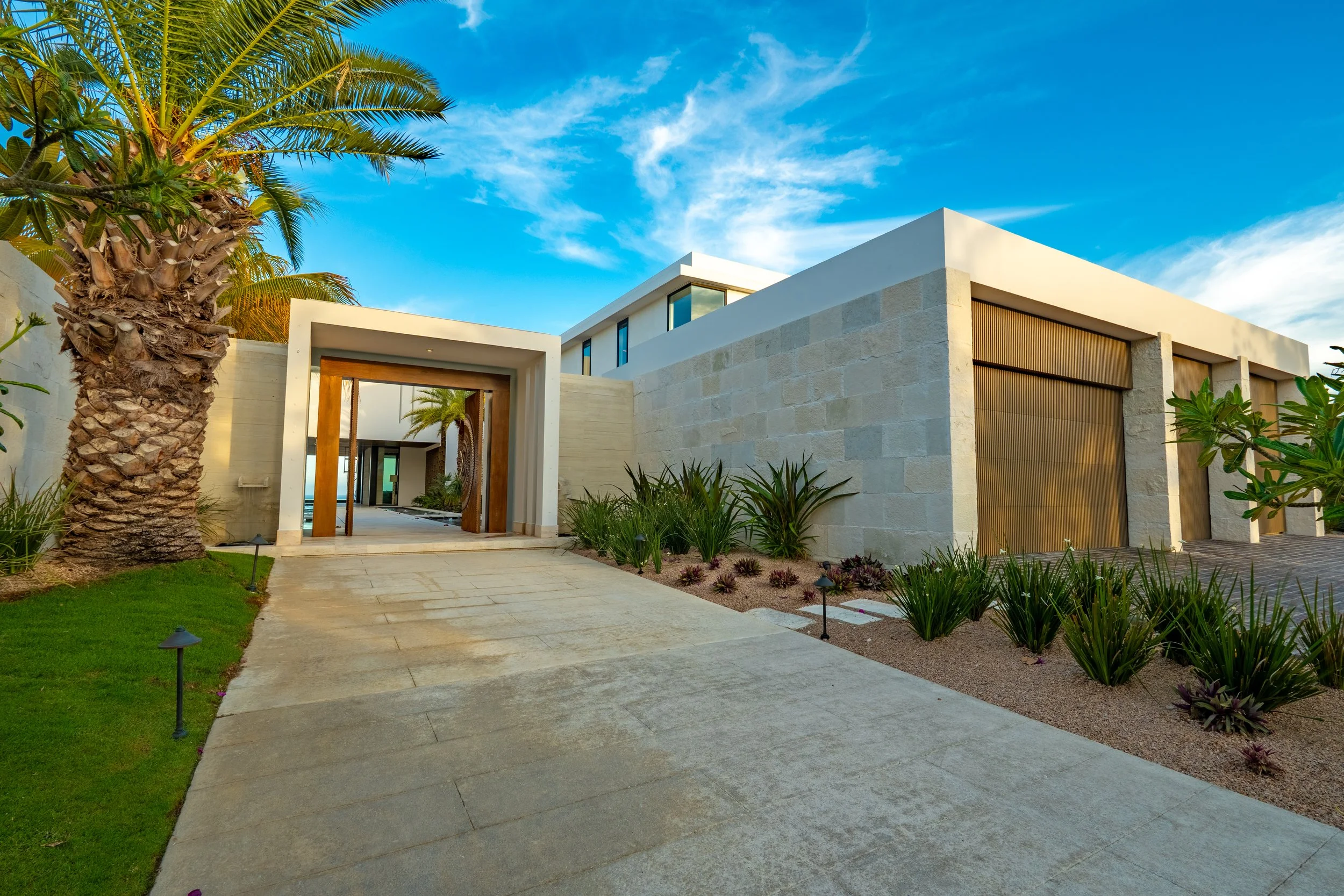 Modern white house with large windows and wooden accents, surrounded by lush green plants and palm trees, under a blue sky with scattered clouds.