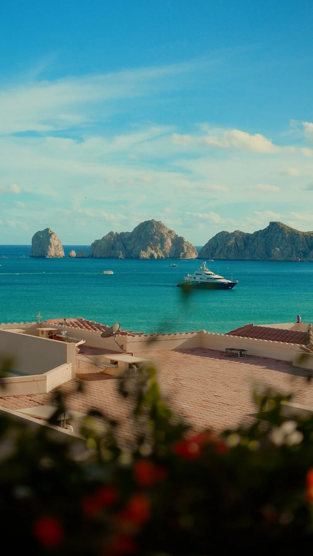 View of a coastal scene from Cabo San Lucas with a yacht in turquoise waters and rocky islands in the background, seen from a terrace with rooftops in the foreground.