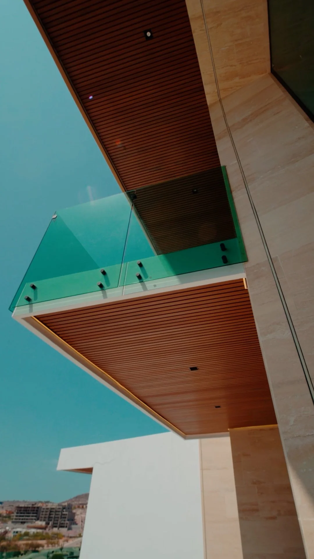 Modern building with a glass balcony, wooden panels, and beige stone walls, under a clear blue sky.