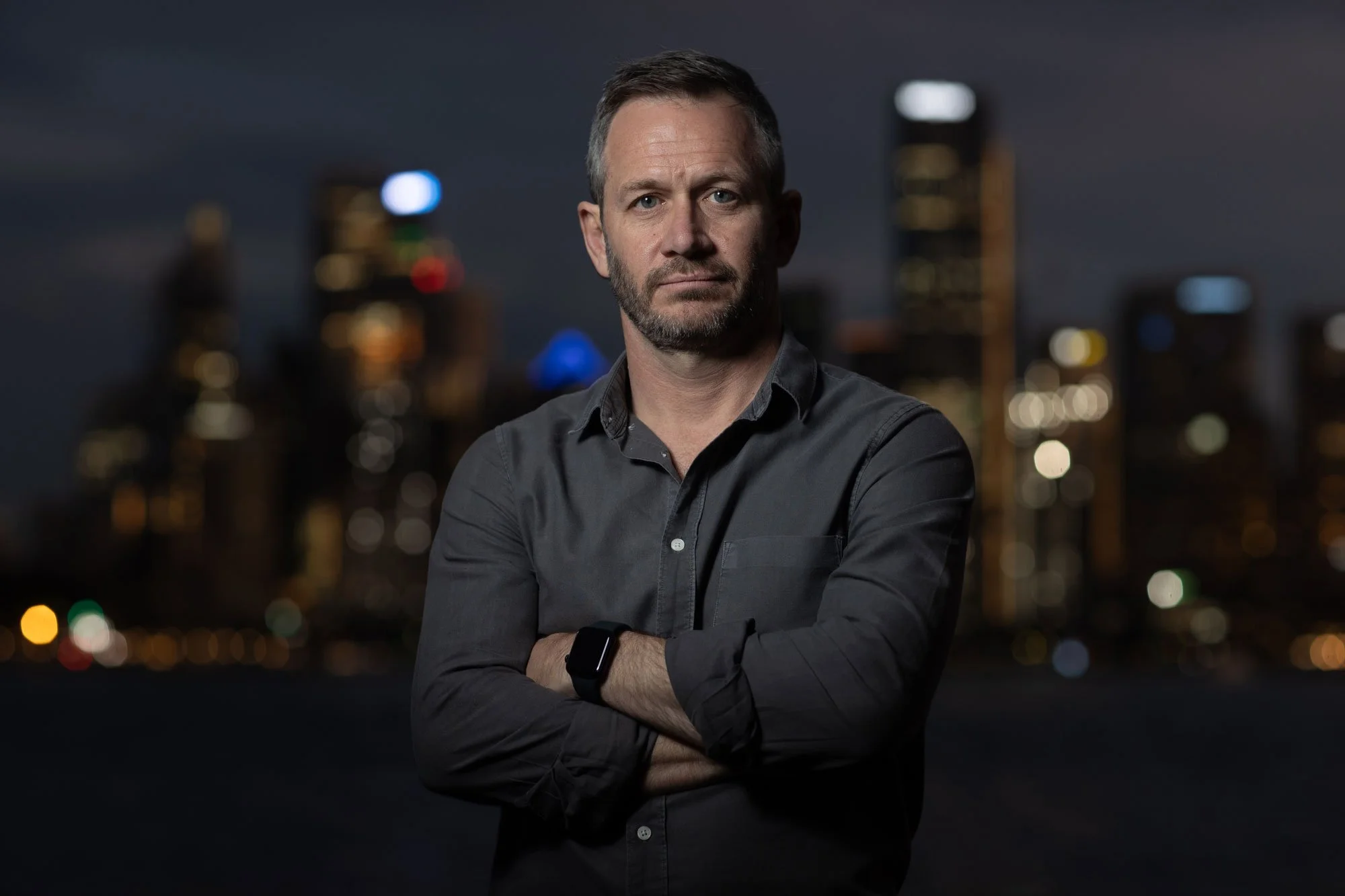 A man with short dark hair and a beard wearing a dark shirt stands with crossed arms in front of the Sydney cityscape at night, with blurred colourful lights in the background.