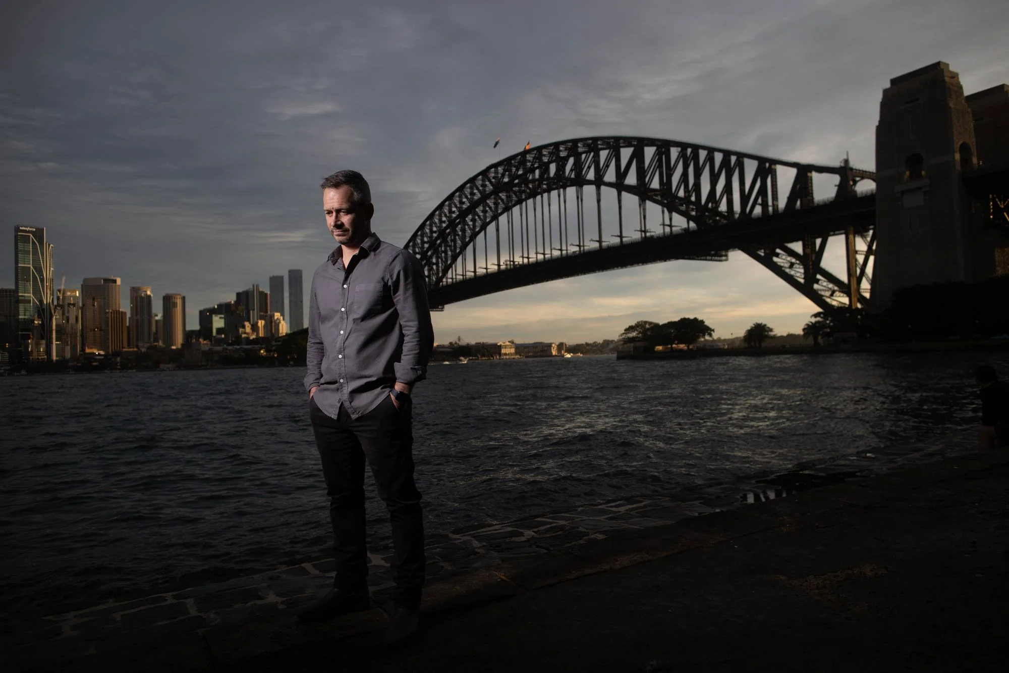 A man stands with his hands in his pockets looking down. He is by the water with the Sydney Harbour Bridge and the Sydney city skyline in the background during dusk.