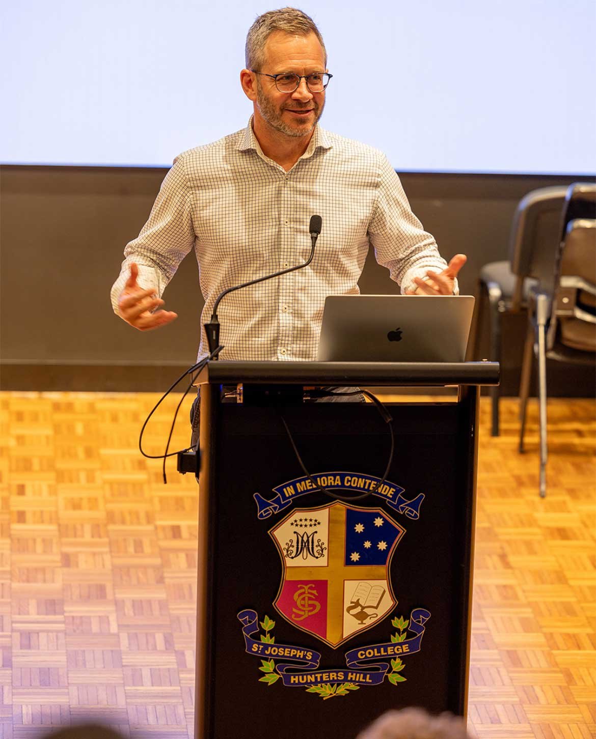 A man with glasses and light hair wearing a checkered shirt speaking at a podium with a microphone, in front of a laptop, at a school. The podium has an emblem with the college's name and crest.