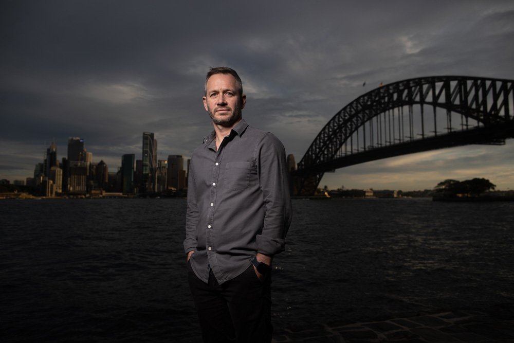 A man in a gray shirt standing by the water with the Sydney city skyline and the Sydney Harbour Bridge in the background during cloudy weather.
