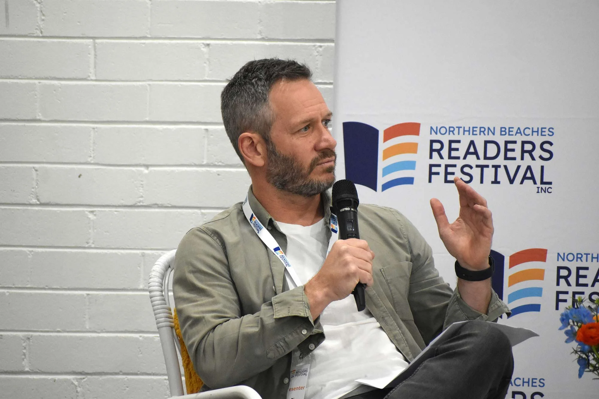 A man with short dark hair and a beard is seated and speaking into a microphone. He is wearing a beige jacket over a white shirt and has a event badge around his neck. Behind him, a banner displays the logo and name for the Northern Beaches Readers F