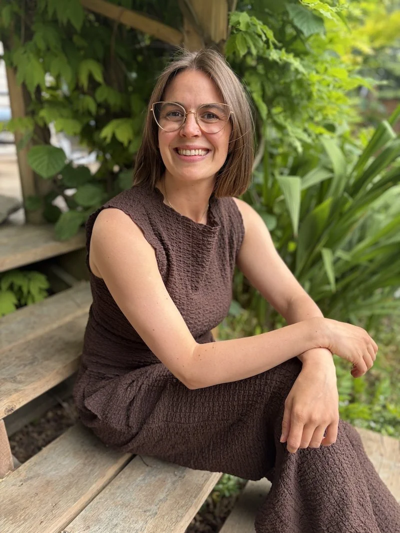 A woman with glasses and shoulder-length hair sitting on wooden steps outdoors, smiling, with green plants and leaves in the background.