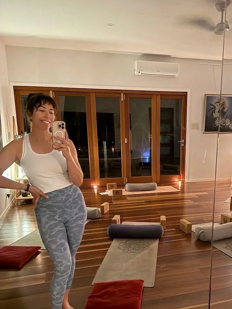 A woman taking a selfie in a room with wooden floors and yoga mats, blocks, and bolsters arranged for a yoga or meditation session.