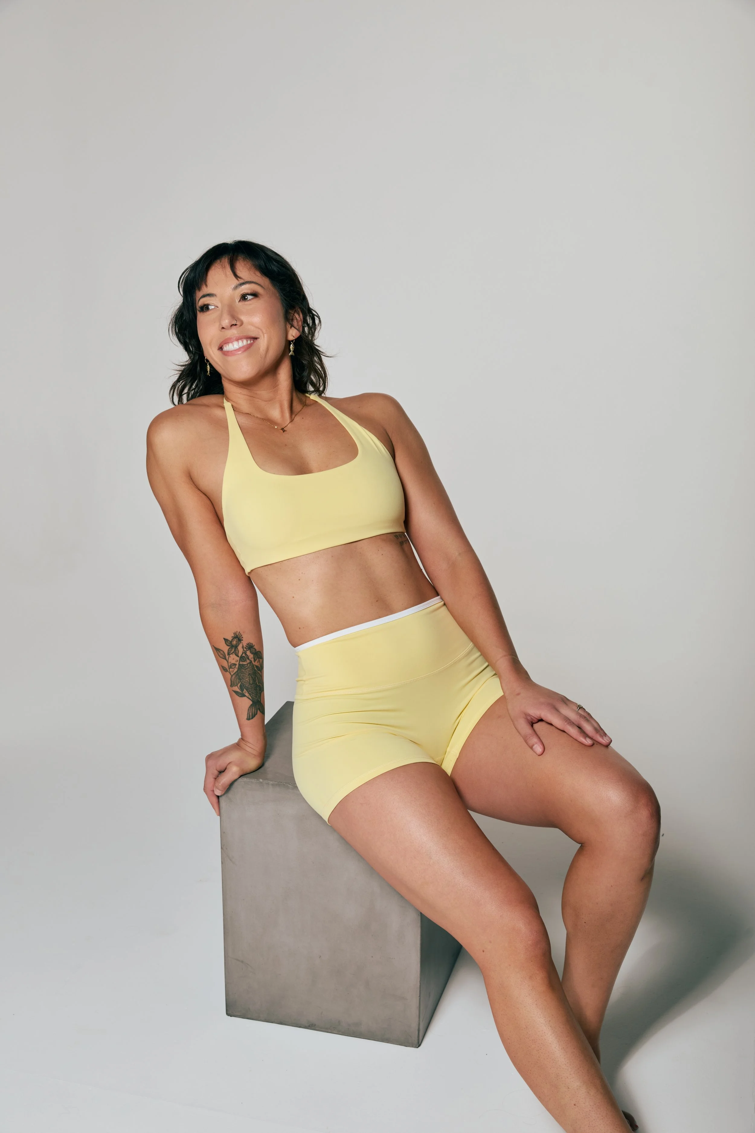 Woman with dark hair and tattoos sitting on a concrete block, wearing yellow sports bra and shorts, smiling and looking to the side in a studio setting with a plain light background.