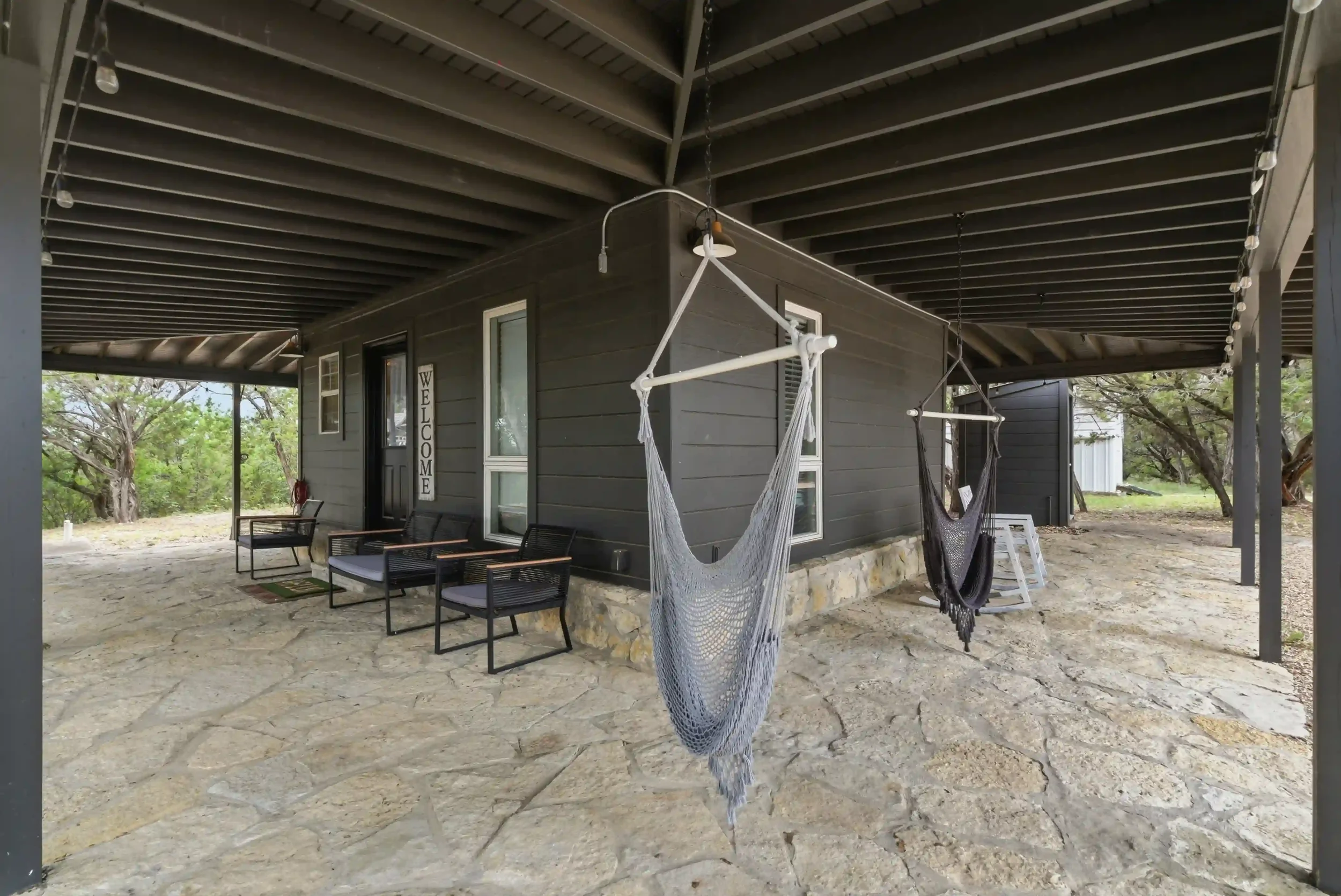 Covered outdoor porch area with hanging hammock chairs, patio seating, and a stone floor with a house in the background and trees beyond.
