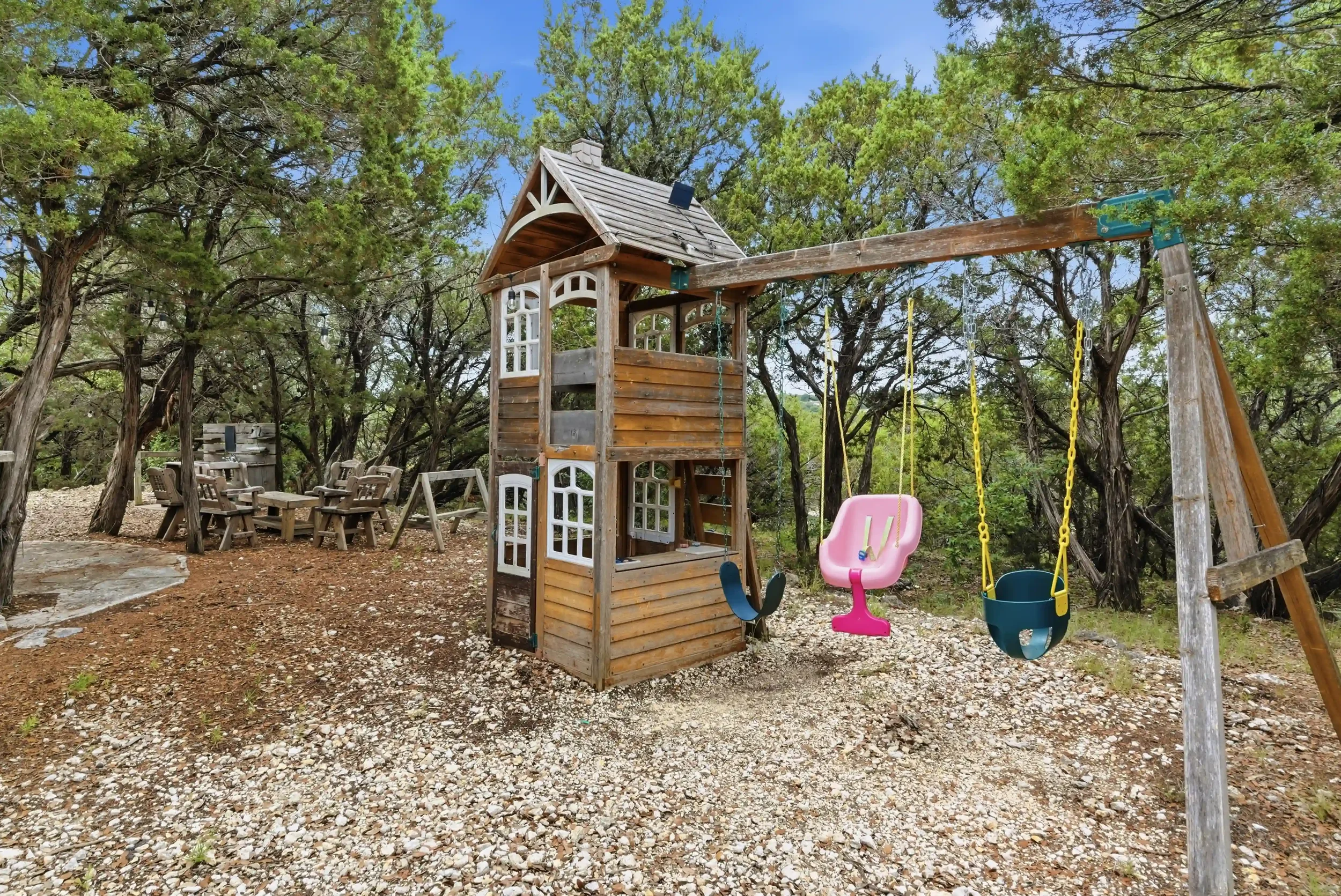 Outdoor play area with wooden playhouse, swings, and a seating area surrounded by trees.
