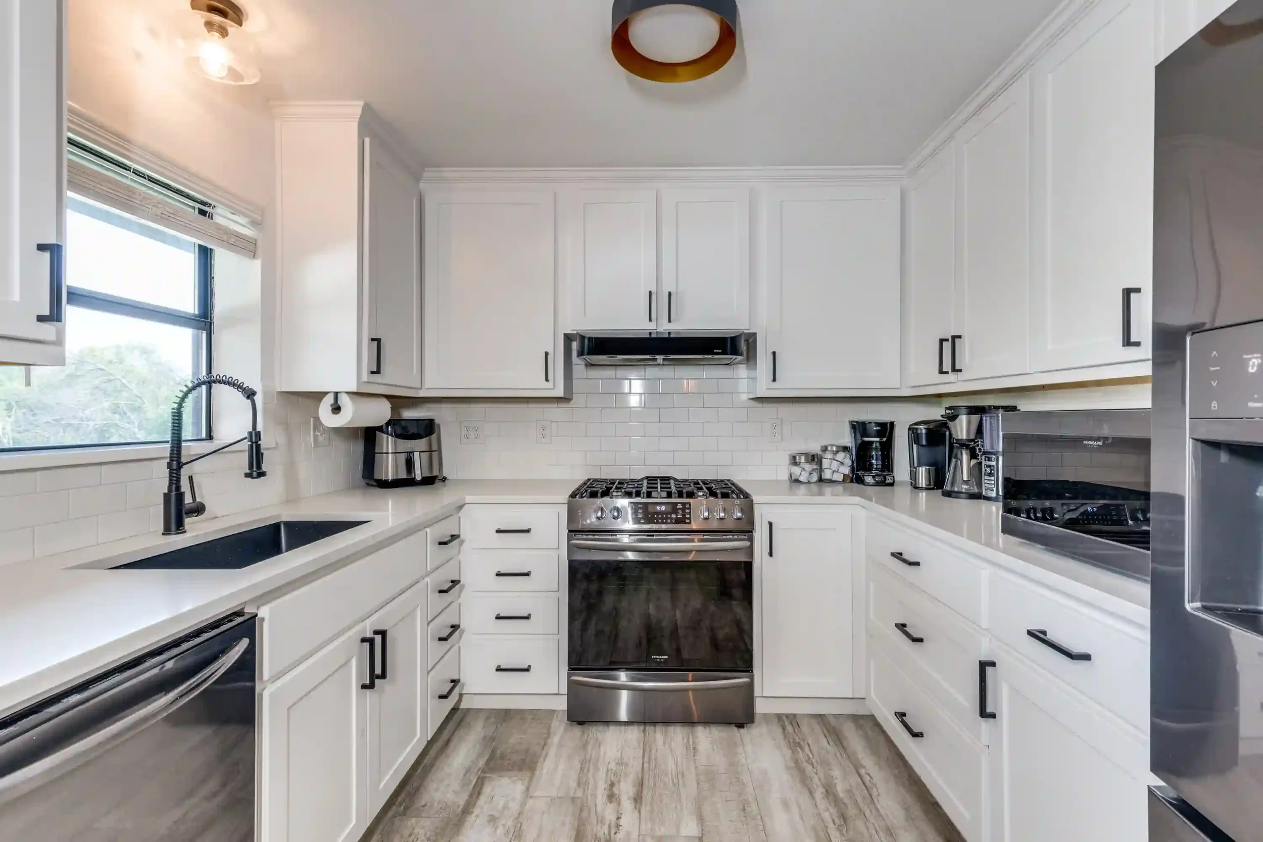 Modern white kitchen with black handles, stainless steel appliances, and a window above the sink, featuring white subway tiles and wooden flooring.