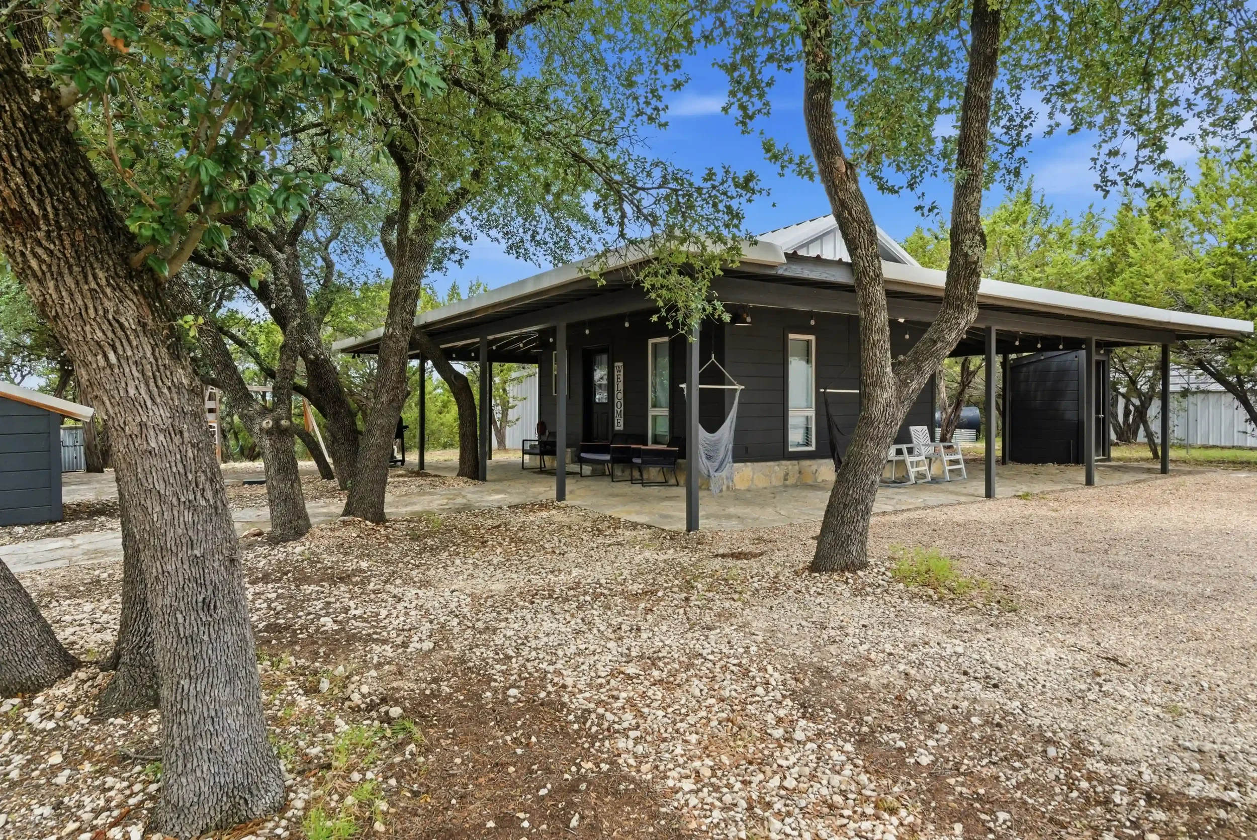 A cozy black house with a porch, surrounded by trees with partial shade, sitting on a gravel yard under a blue sky.